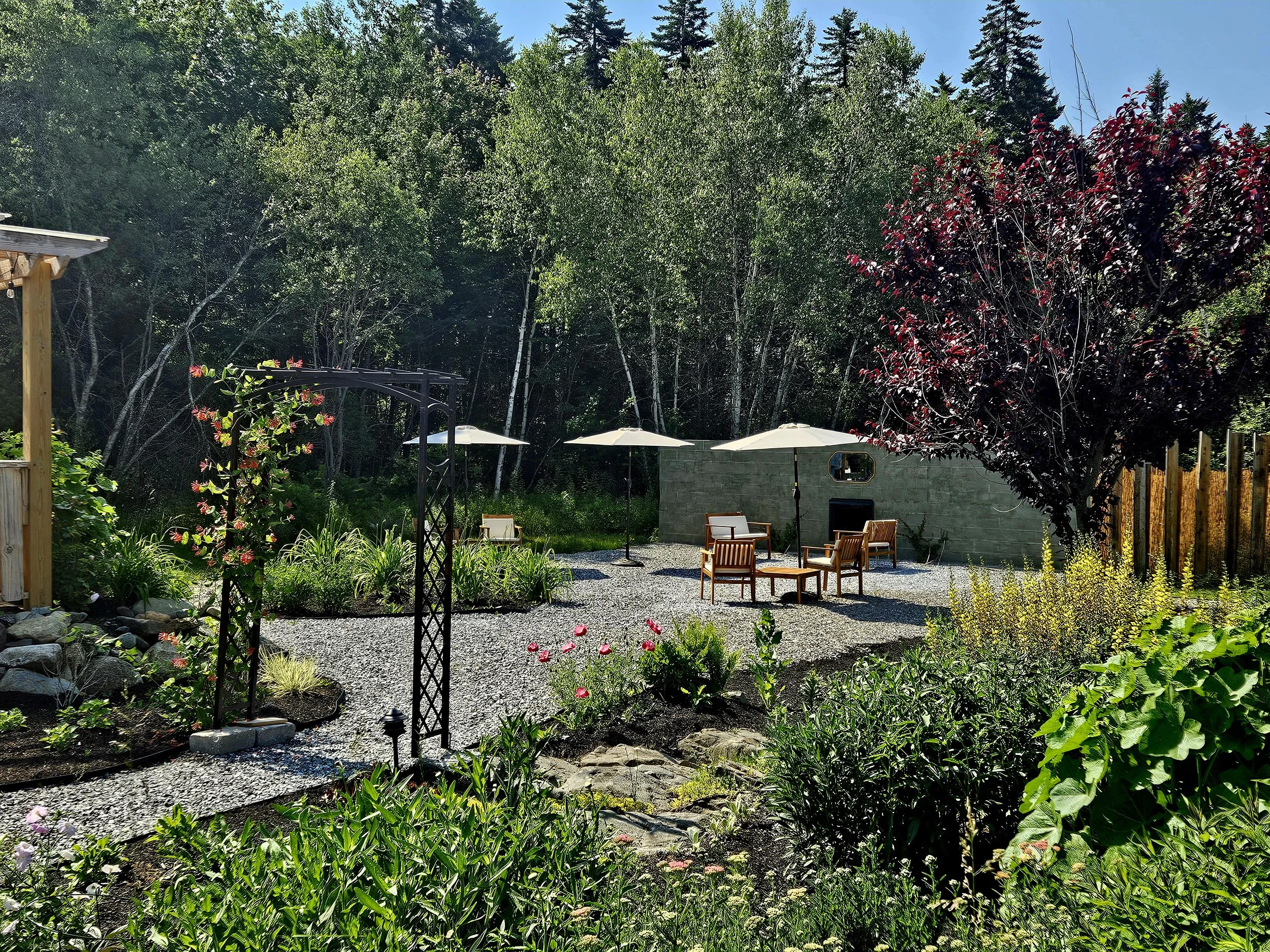 A backyard garden patio with walking paths, garden beds, outdoor furniture, and umbrellas surrounded by lush trees and plants.