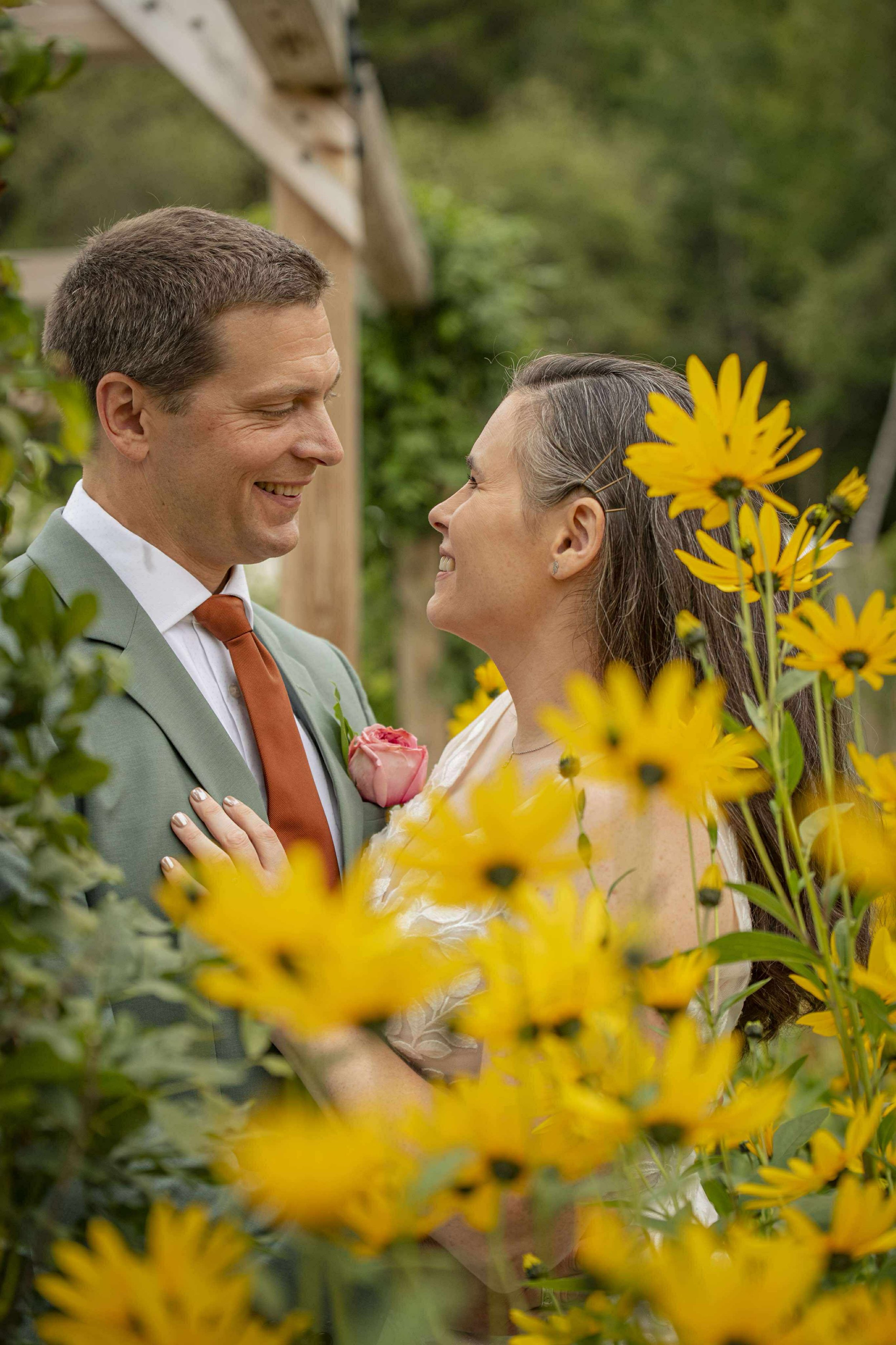 A couple dressed in wedding attire smiling at each other among yellow flowers in a garden setting.