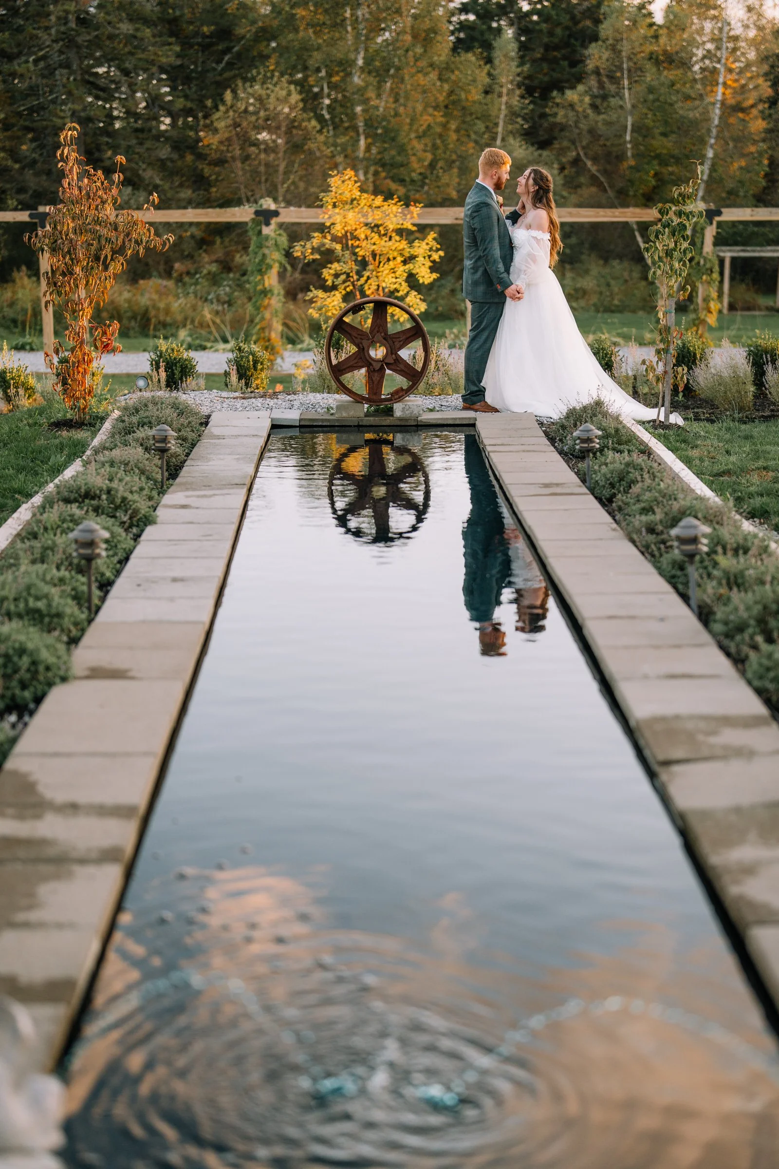 A bride and groom standing face-to-face holding hands at their wedding, in an outdoor setting with autumn trees and a small reflective pond in front of them.