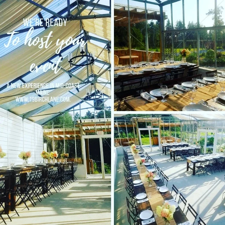 Indoor event space with glass walls and ceiling, decorated for a celebration or gathering. Two long tables are set with flowers, plates, glasses, and silverware, with sunlight streaming in and a view of greenery outside.