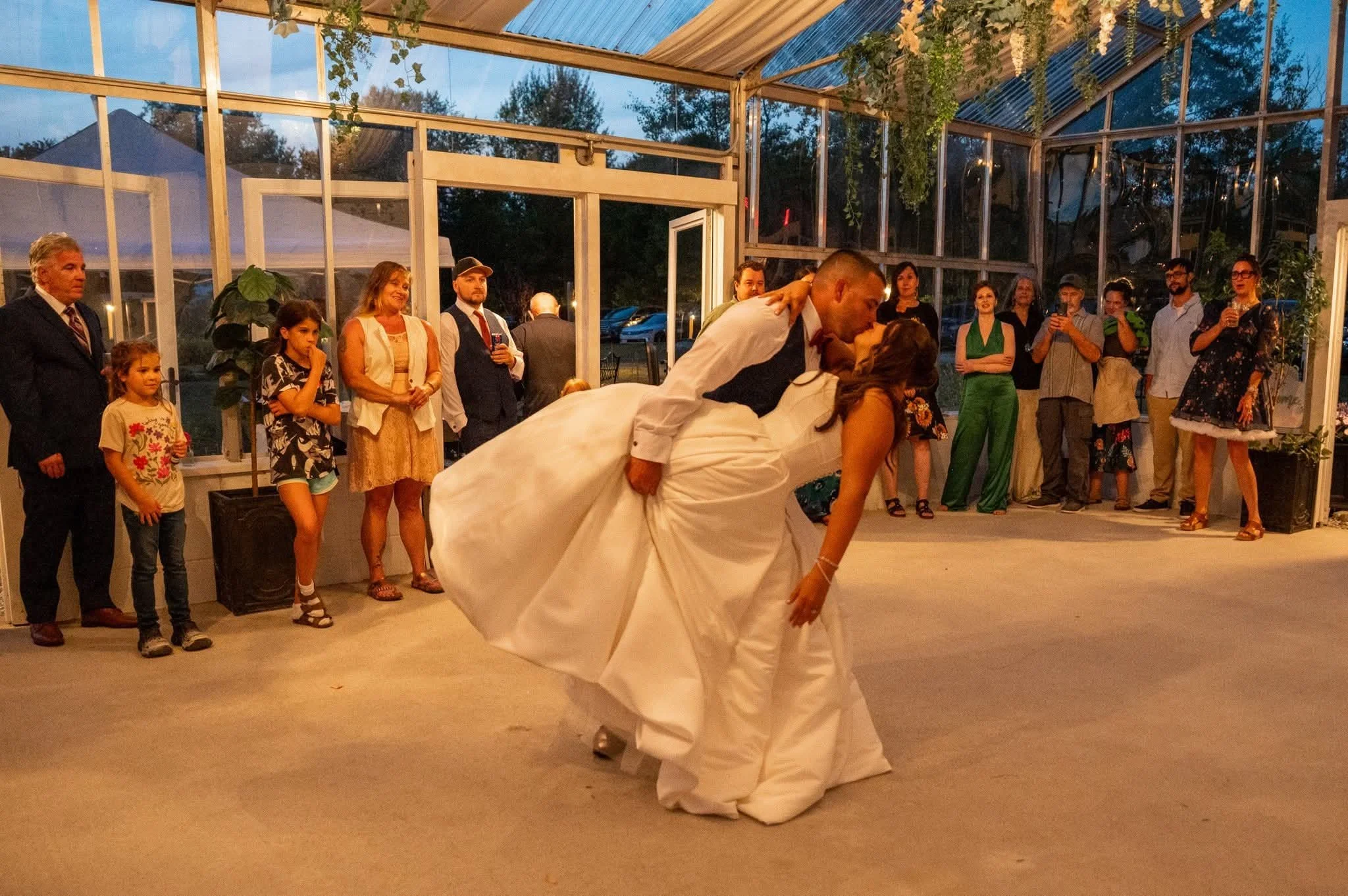 A bride and groom kiss during their wedding reception as guests gather around in a glass-walled venue at dusk.