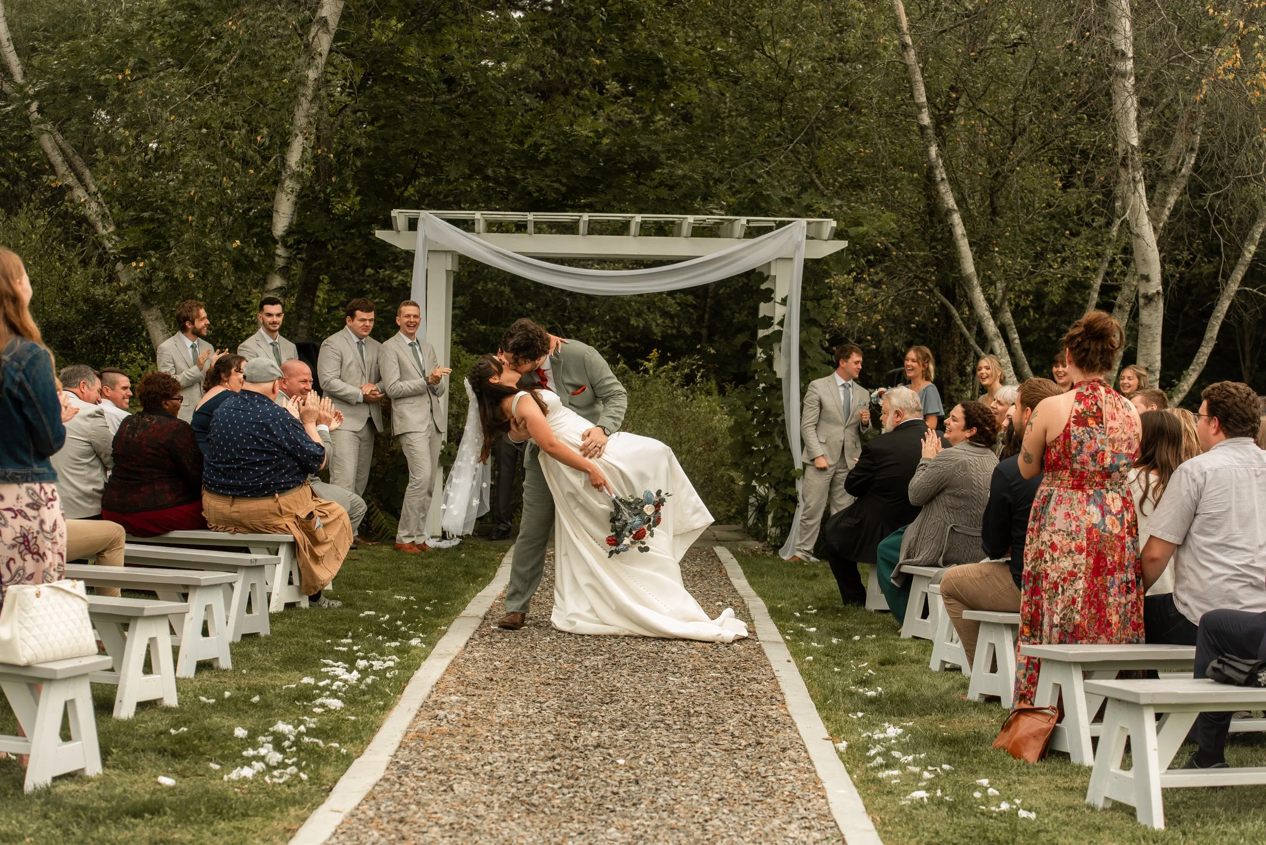 A newlywed couple shares a kiss during their outdoor wedding ceremony, with guests seated on benches and a decorated arch surrounded by trees in the background.