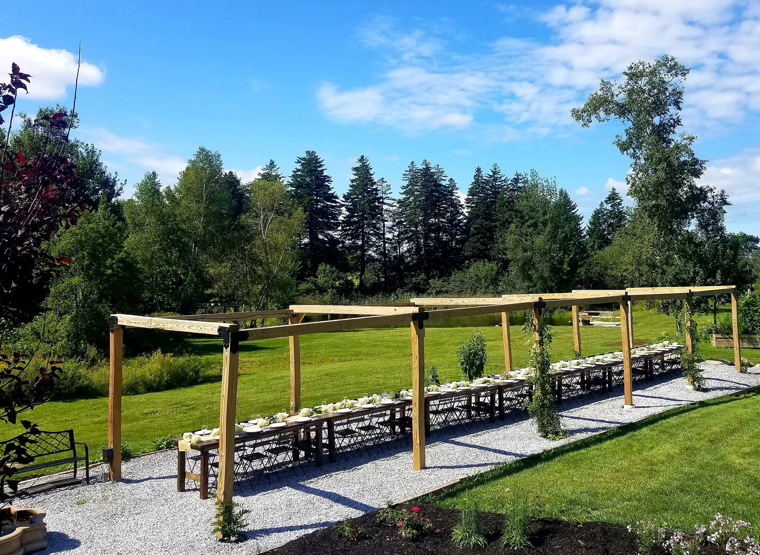 Outdoor dining setup with a long table, chairs, and a wooden framework, set in a grassy area with trees and a blue sky in the background.