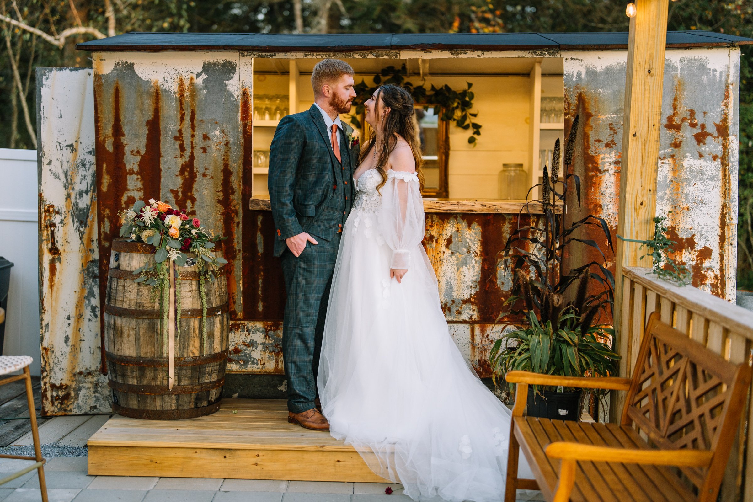 A bride and groom stand close together during their wedding, gazing into each other's eyes. The groom is wearing a green plaid suit and the bride is in a white wedding dress with off-the-shoulder sleeves. They are indoors in front of a rustic, weathe