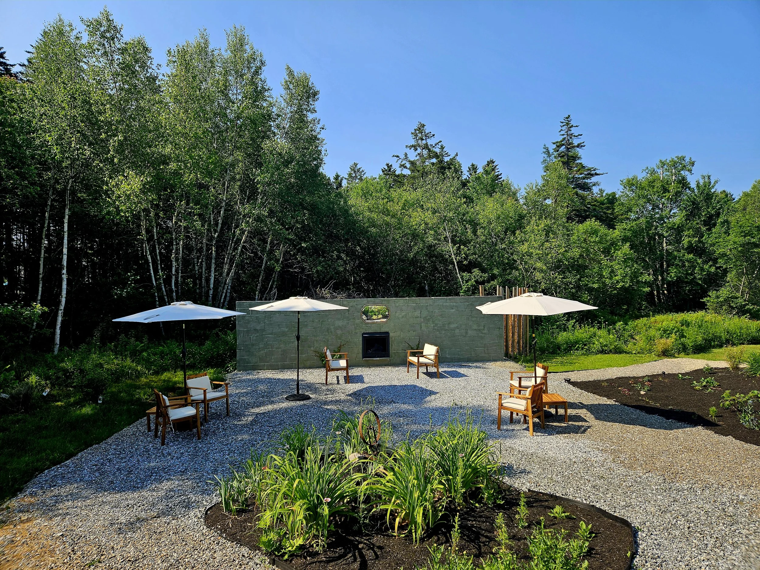 A backyard patio with four chairs, three umbrellas, a green wall with a built-in fireplace, and lush green trees in the background under a clear blue sky.