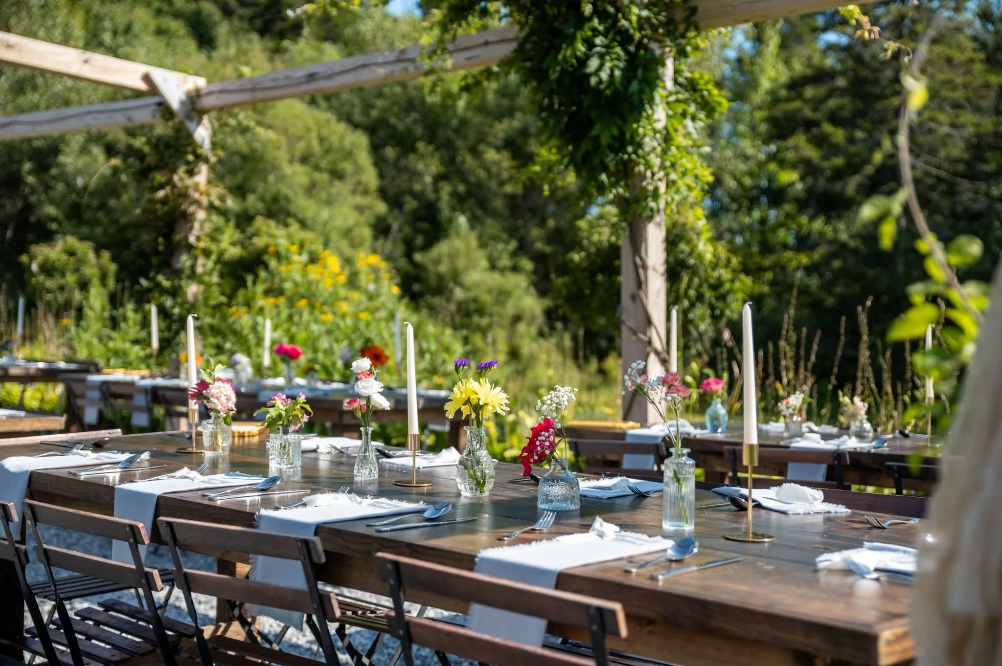 An outdoor dining table decorated with flowers and candles, set for a meal in a garden with lush green trees and plants in the background.