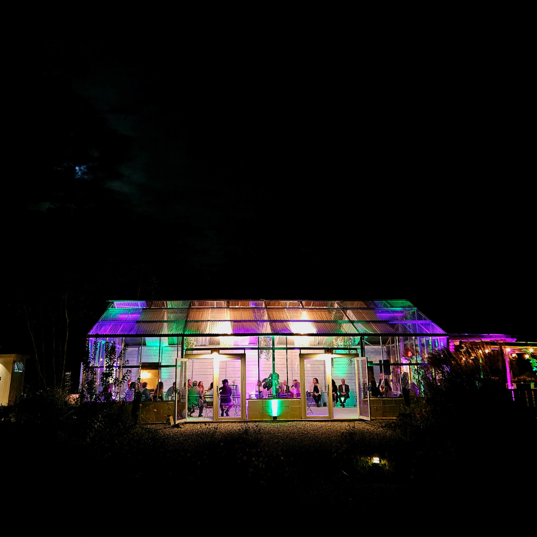 Nighttime scene of a glass greenhouse or conservatory with colorful purple, green, and orange lighting, with people inside dining or socializing, set against a dark sky.
