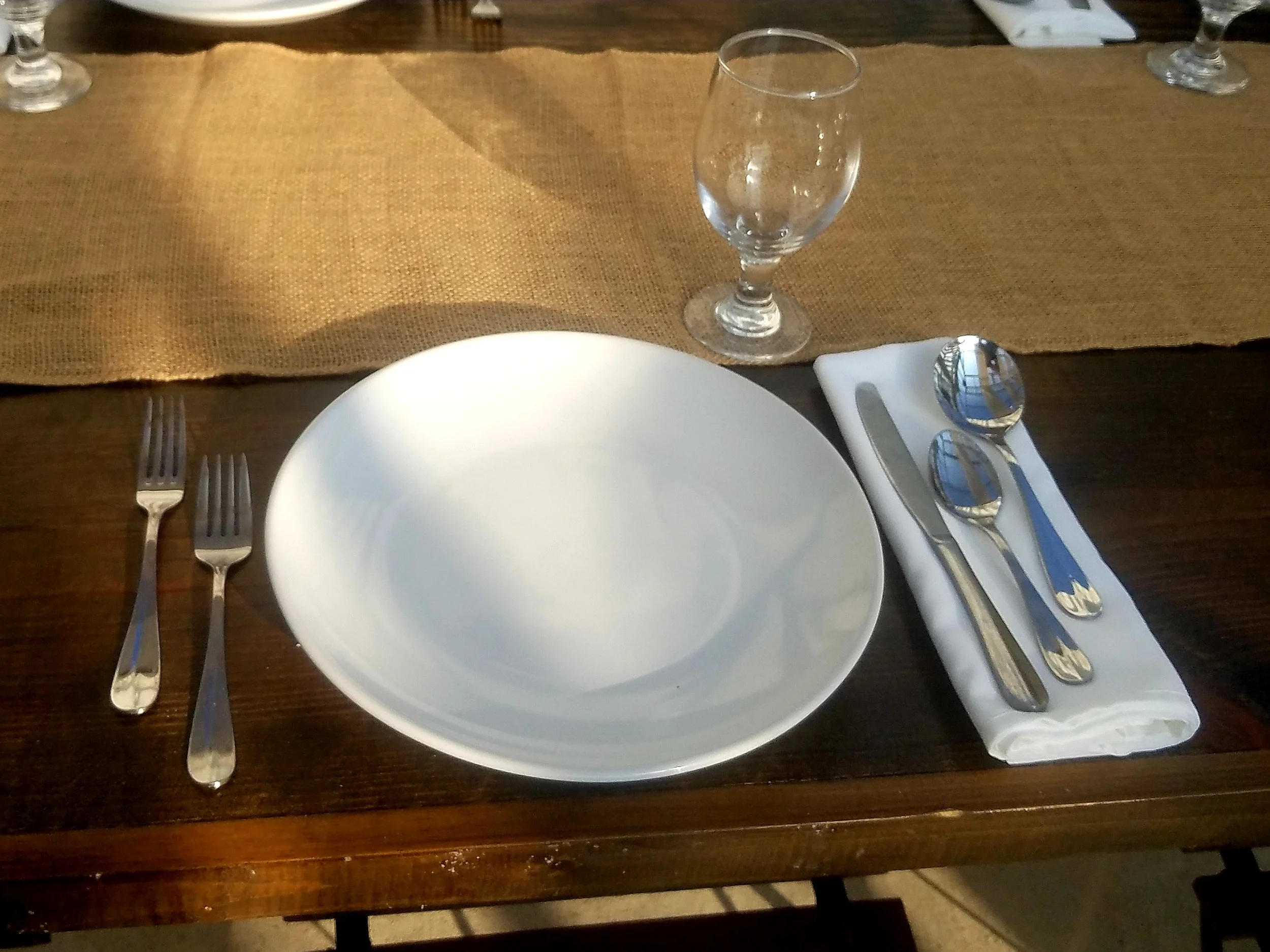 Empty white plate on dark wooden table with silverware and a glass of water.