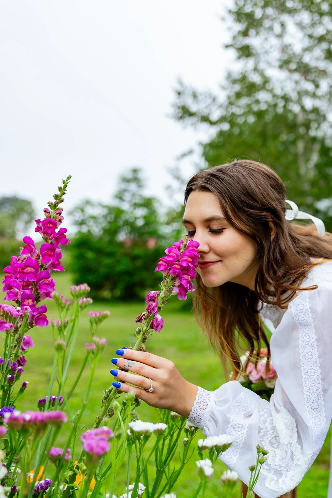 Young woman with brown hair, wearing a white lace dress, smelling pink flowers in a garden.