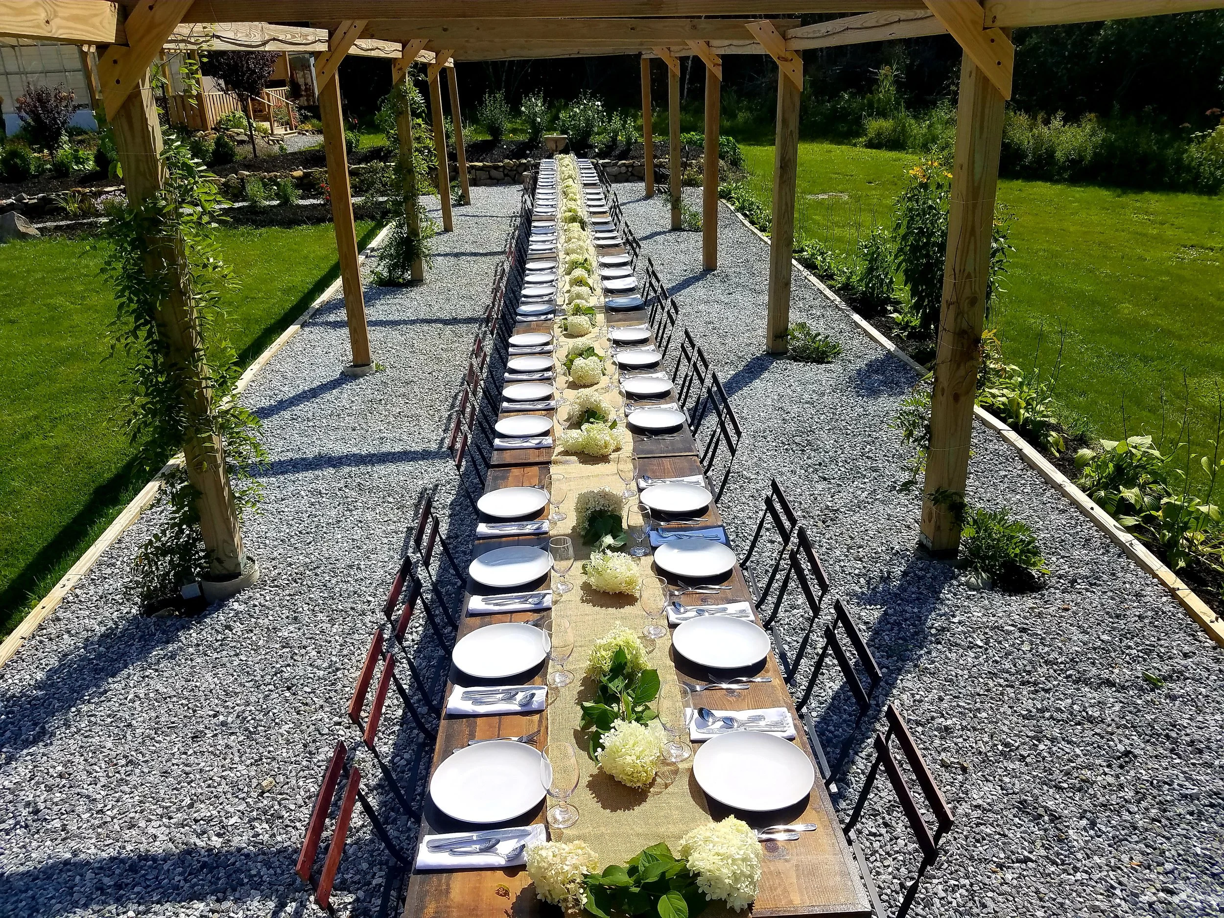 An outdoor dining setup featuring a long wooden table under a wooden pergola with black chairs around it, set with white plates, silverware, and floral centerpieces of white hydrangeas. The area is surrounded by greenery and gravel ground.