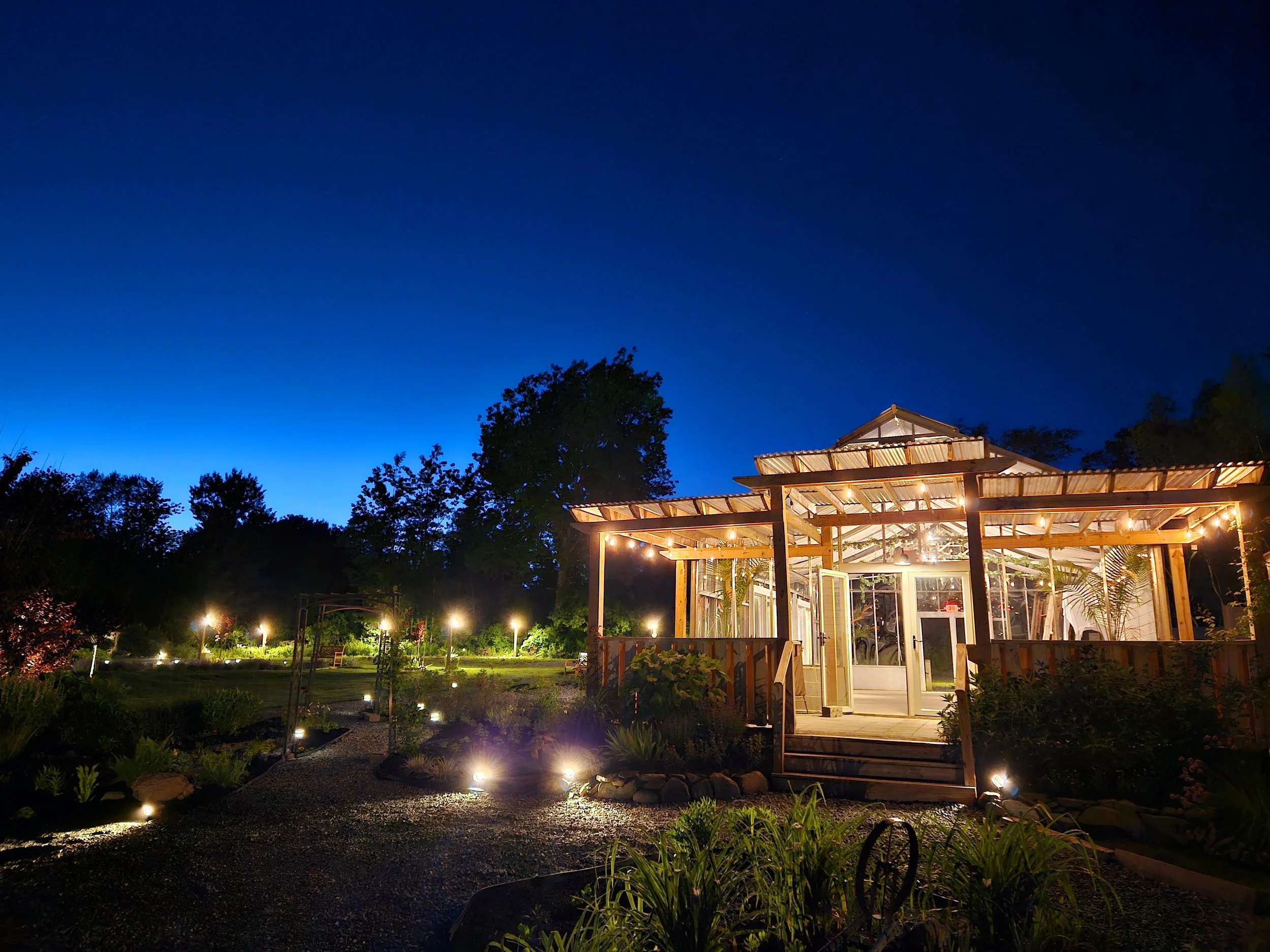 A cozy, well-lit glass house in a garden at night with trees and pathway lights in the background.