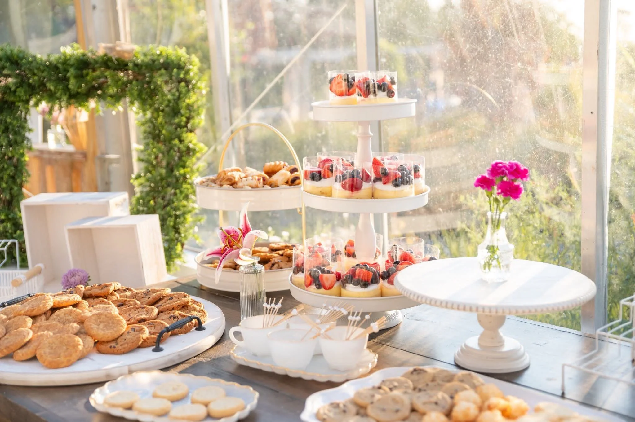 A dessert table with various cookies, fruit tarts, and cupcakes, set in a sunlit greenhouse, decorated with pink flowers.