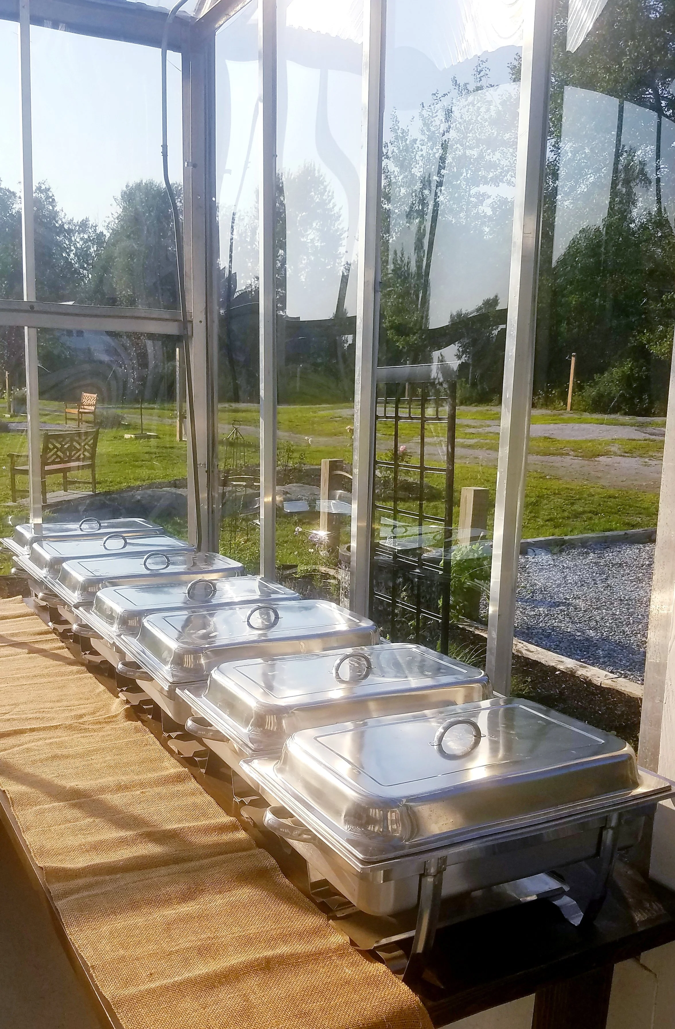 Multiple silver chafing dishes with lids on a table inside a greenhouse, with trees and a grassy area visible outside.