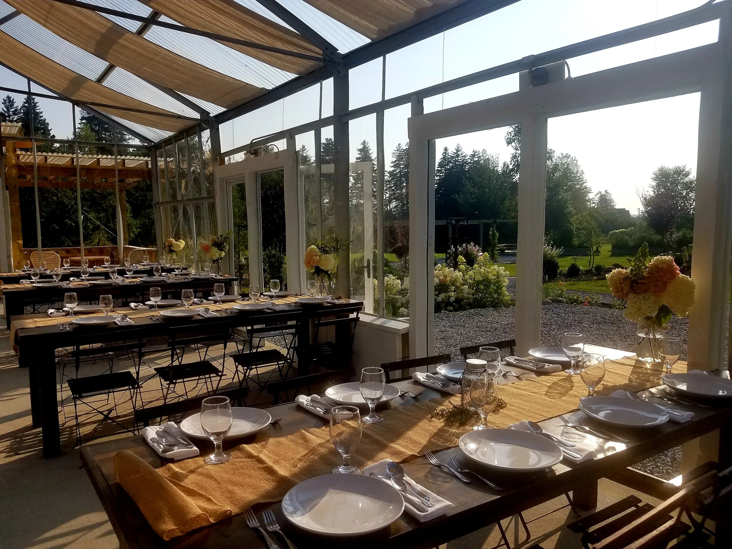 A sunlit glass-enclosed dining area with tables set with white plates, napkins, wine glasses, and floral centerpieces, overlooking a lush garden.