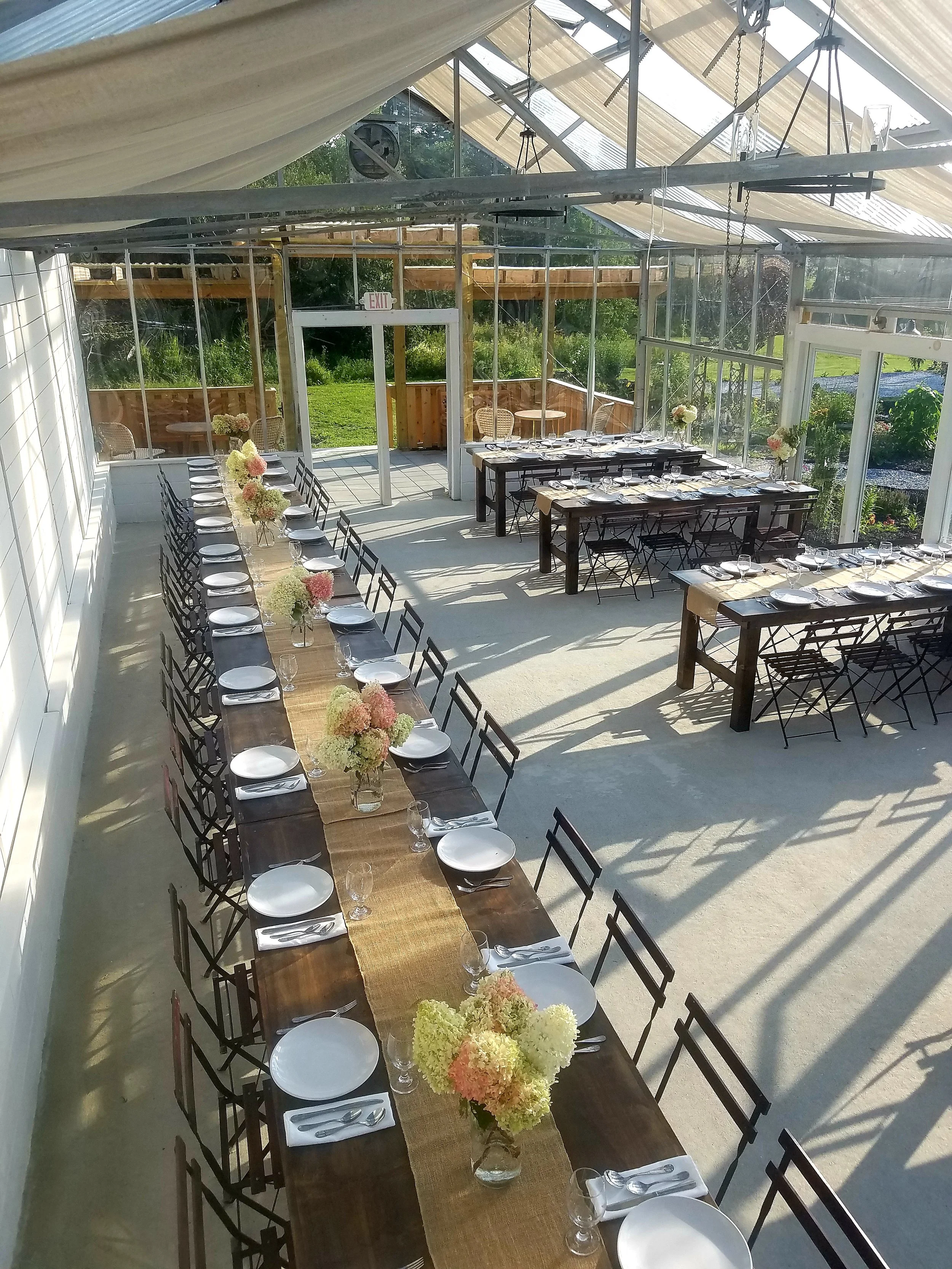 Long dining table set for a wedding or event with white plates, glassware, and napkins, decorated with flower arrangements in vases, in a glass-enclosed venue with views of a grassy yard.
