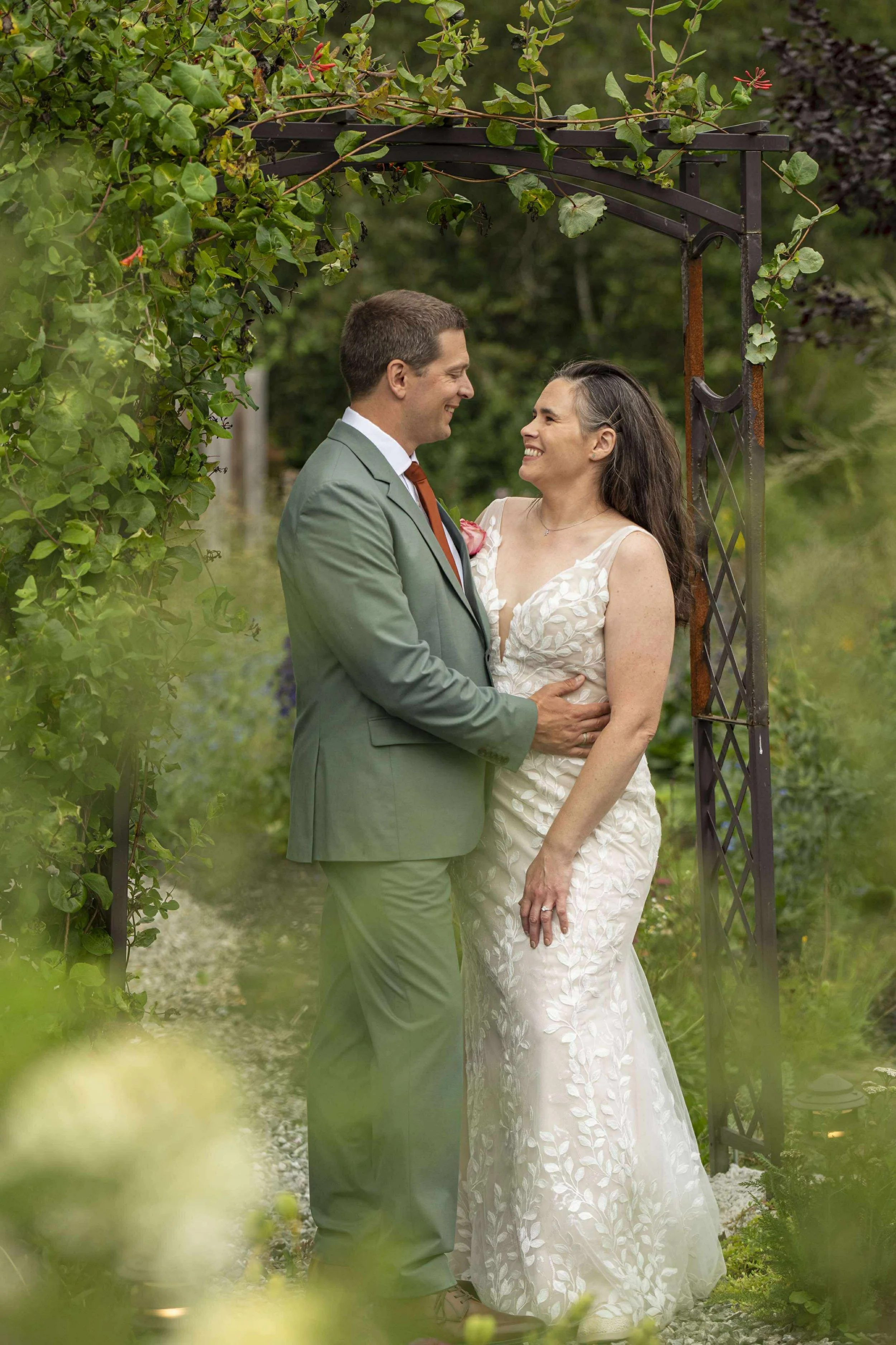 A couple standing under an archway, smiling at each other during a wedding or romantic outdoor event, surrounded by green foliage.
