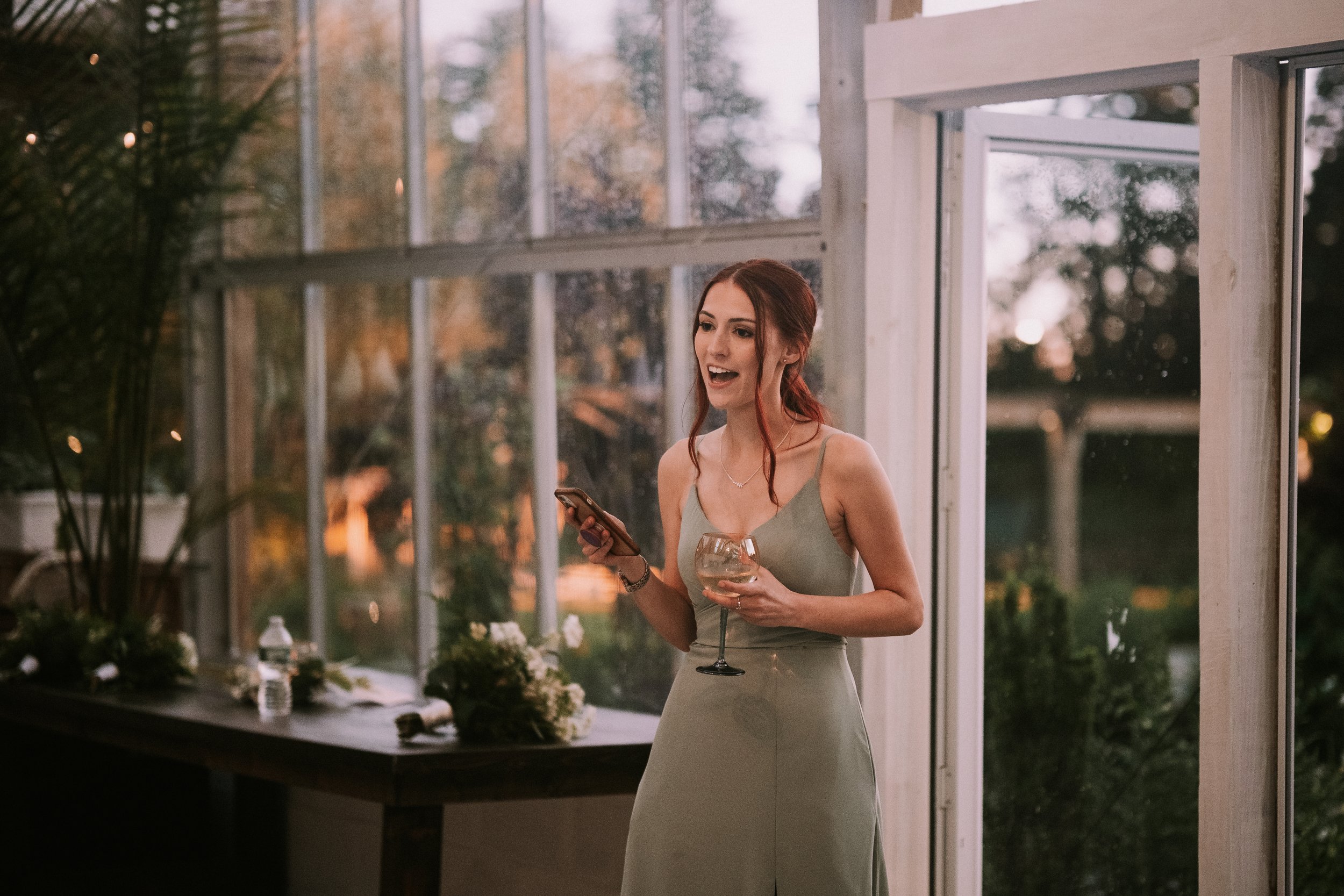 A woman with red hair wearing a green dress, holding a wine glass and a smartphone, speaking at an indoor event in a sunlit room with large windows and greenery outside.