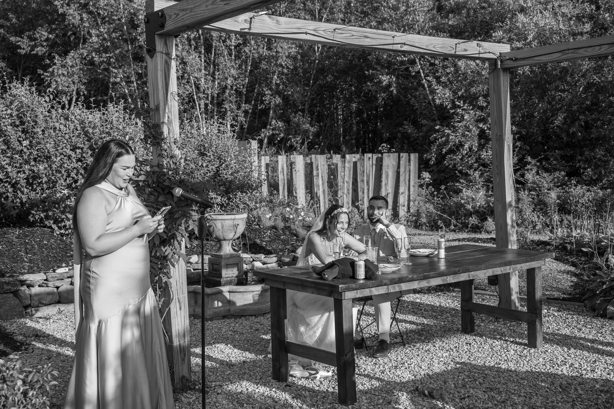 A bride and groom seated at a wooden table during an outdoor wedding reception, with a woman giving a speech or toast next to them and a garden backdrop.