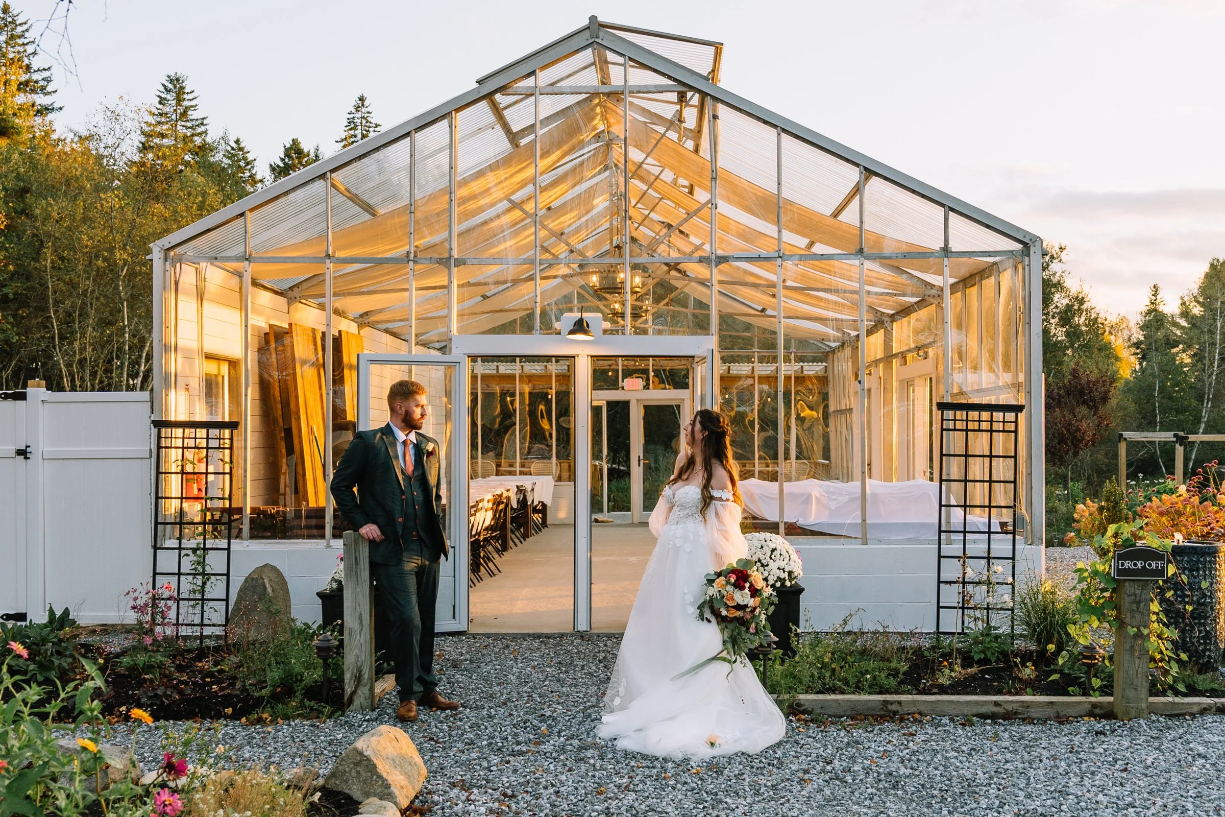 A bride in a white wedding gown holding a bouquet stands outside a transparent greenhouse, talking to a groom in a dark suit, in a garden at sunset.