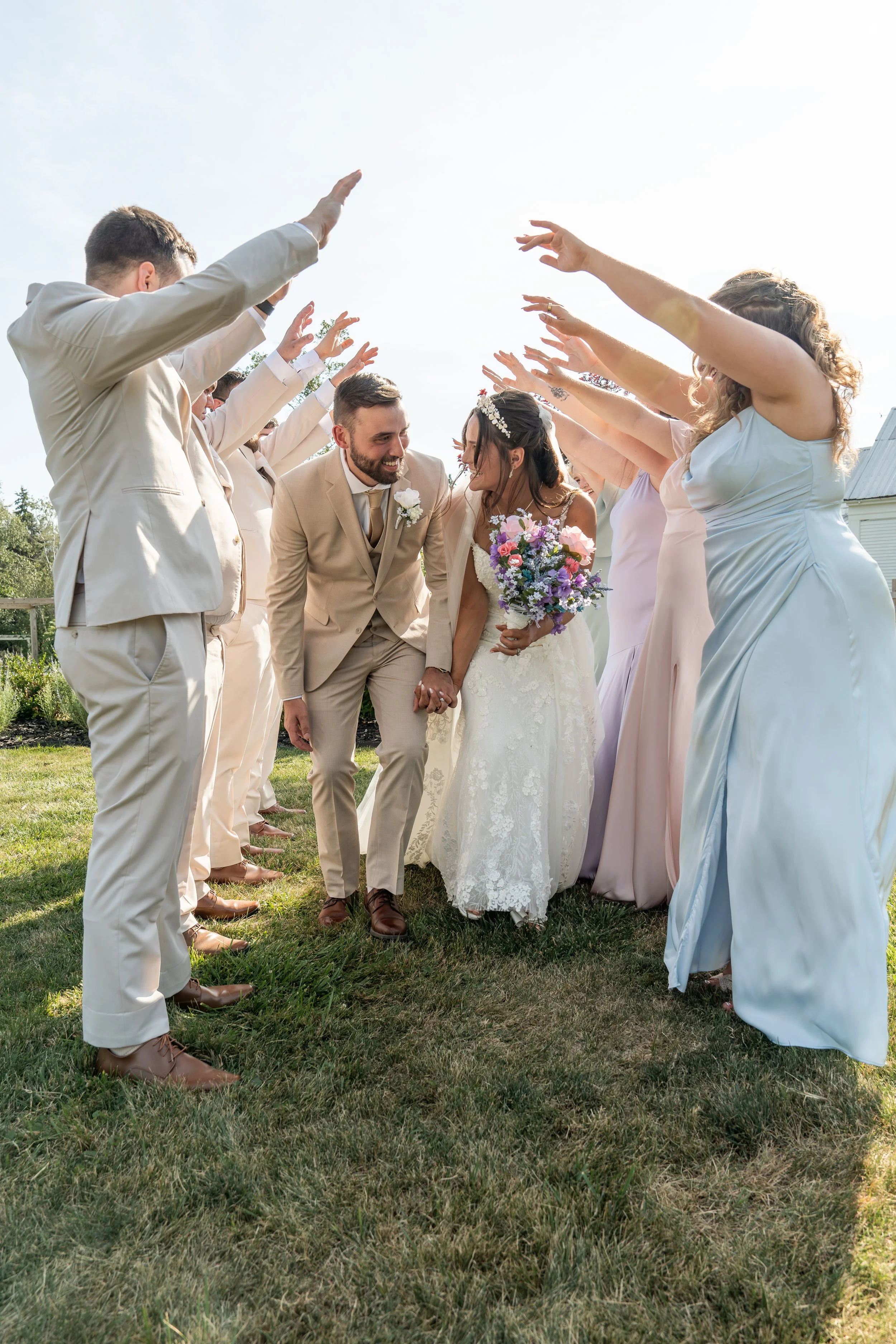 Bride and groom holding hands walking through a tunnel of friends and family with arms raised on a grassy outdoor area during a wedding celebration.