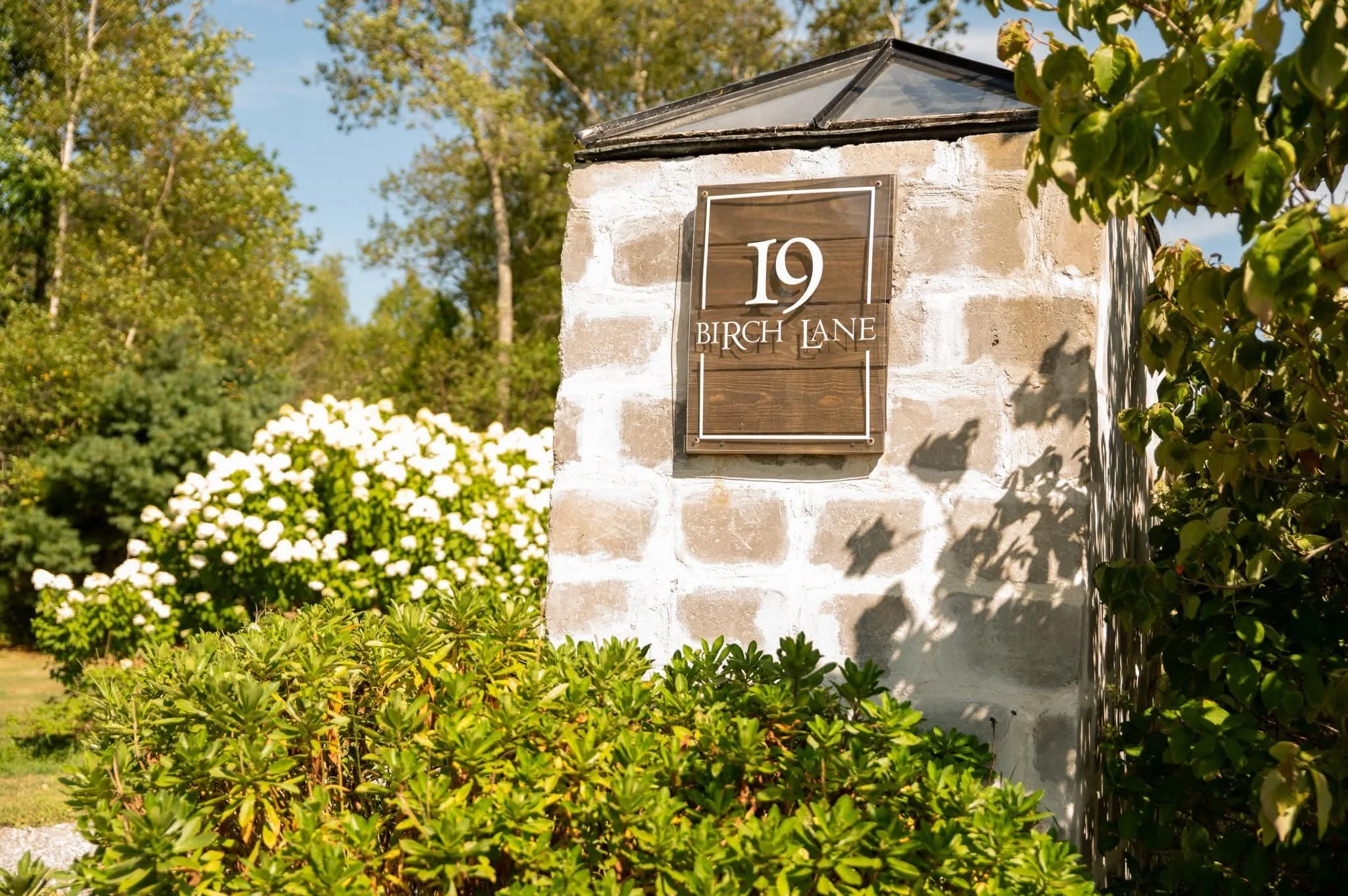 House address sign on brick pillar reading '19 Birch Lane' with greenery and flowering bushes in the background.