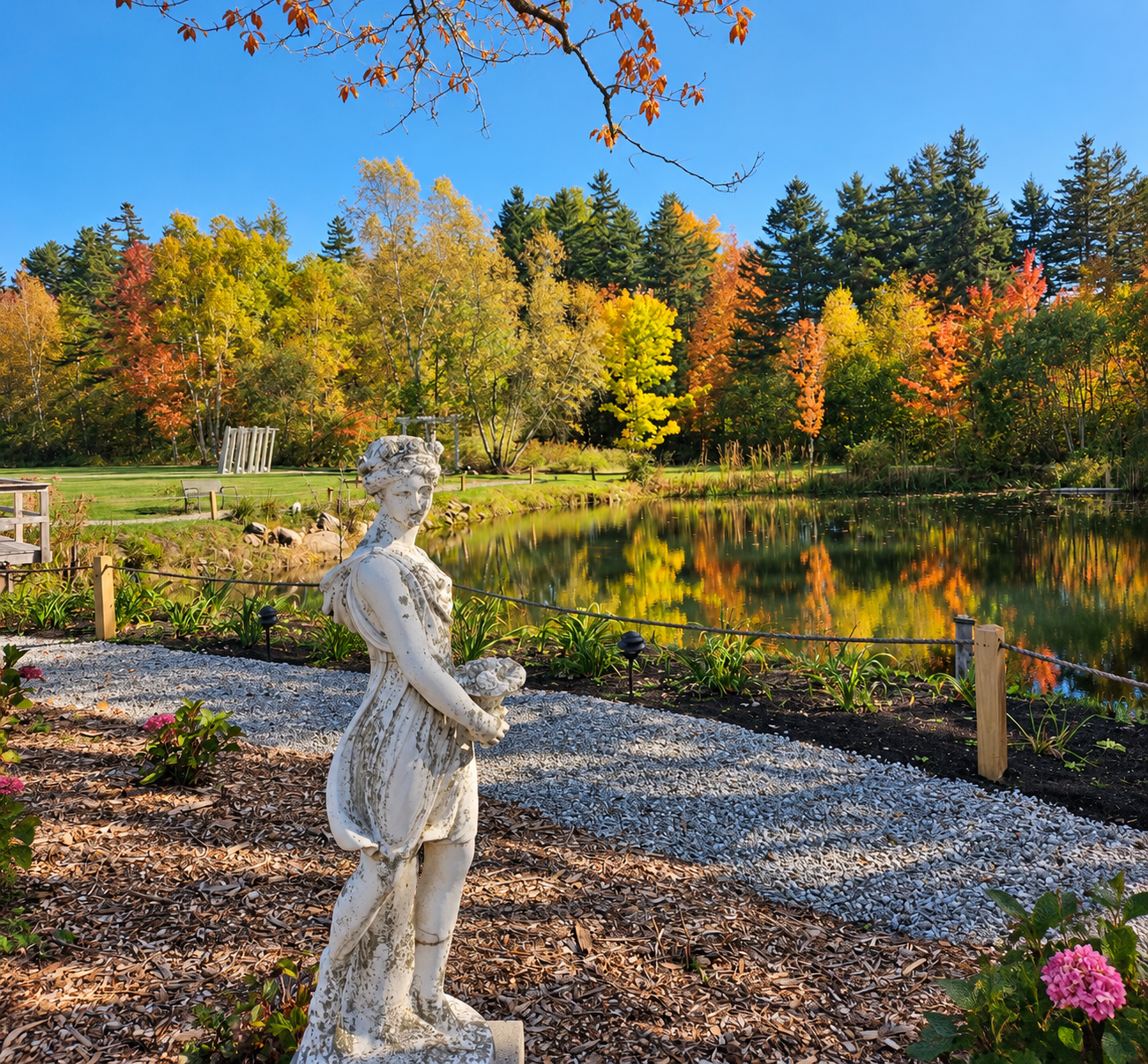 A serene garden scene with a statue of a woman holding a bird, surrounded by colorful trees, a pond reflecting the autumn foliage, and a clear blue sky.