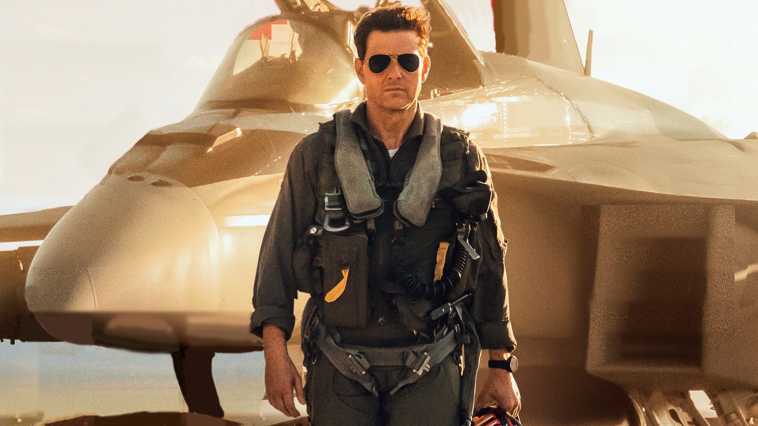 Tom Cruise, wearing a jumpsuit and aviator sunglasses, walking toward the camera with an airplane parked behind him.