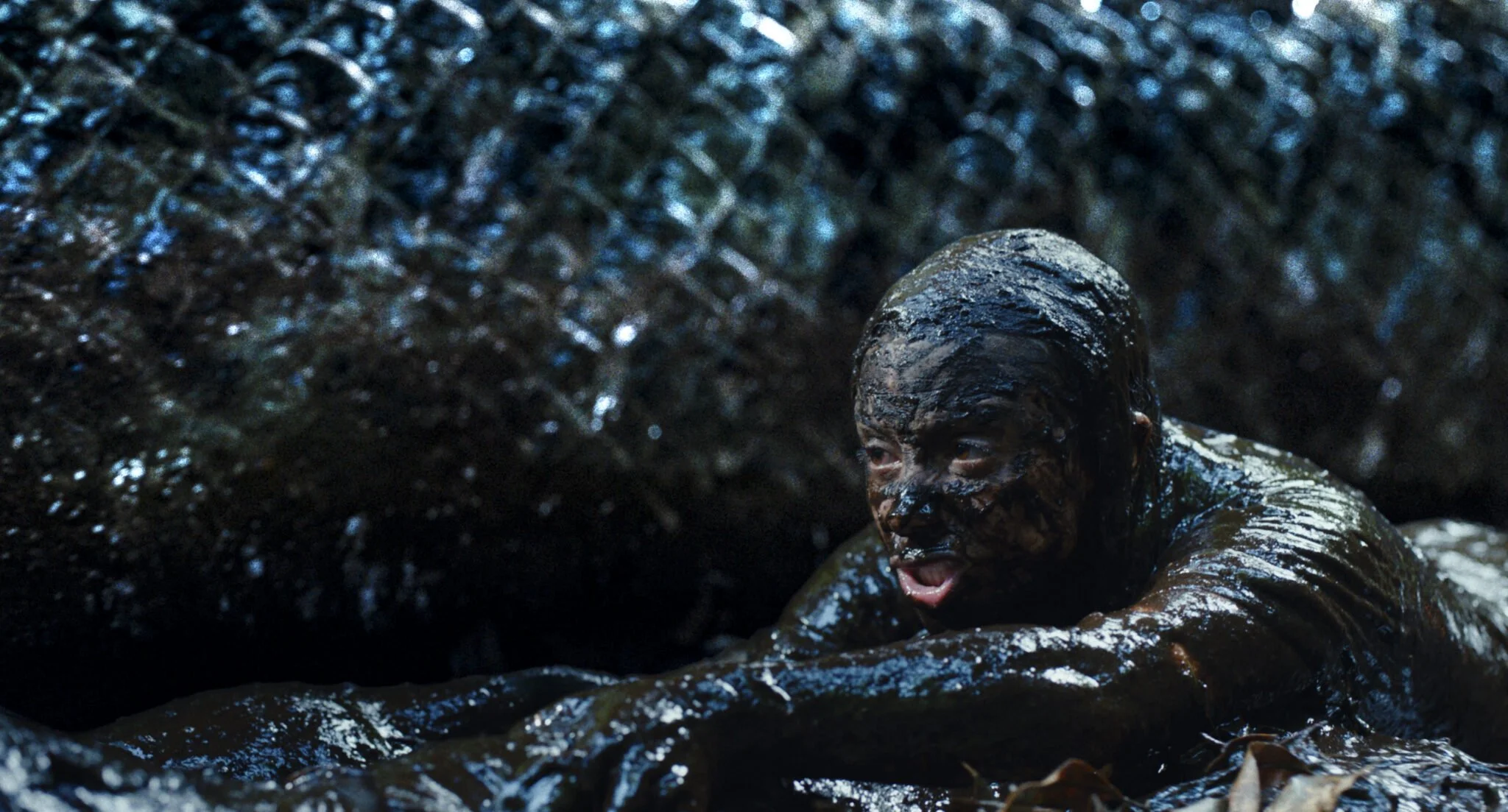 A person, covered in mud, crawling on their stomach in front of a chain link fence at night.