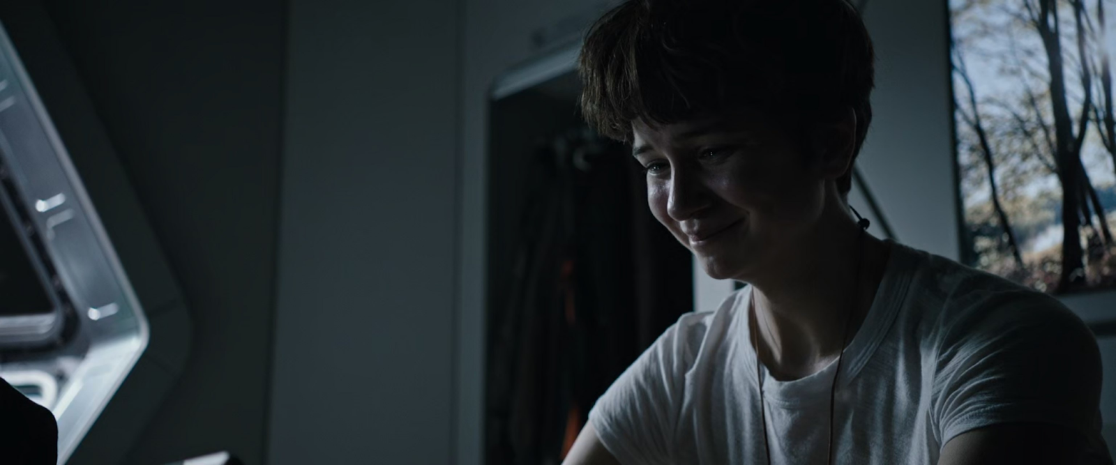 A short-haired person sits in the shadows with faint white light shining on their face. They are grimacing and look upset.