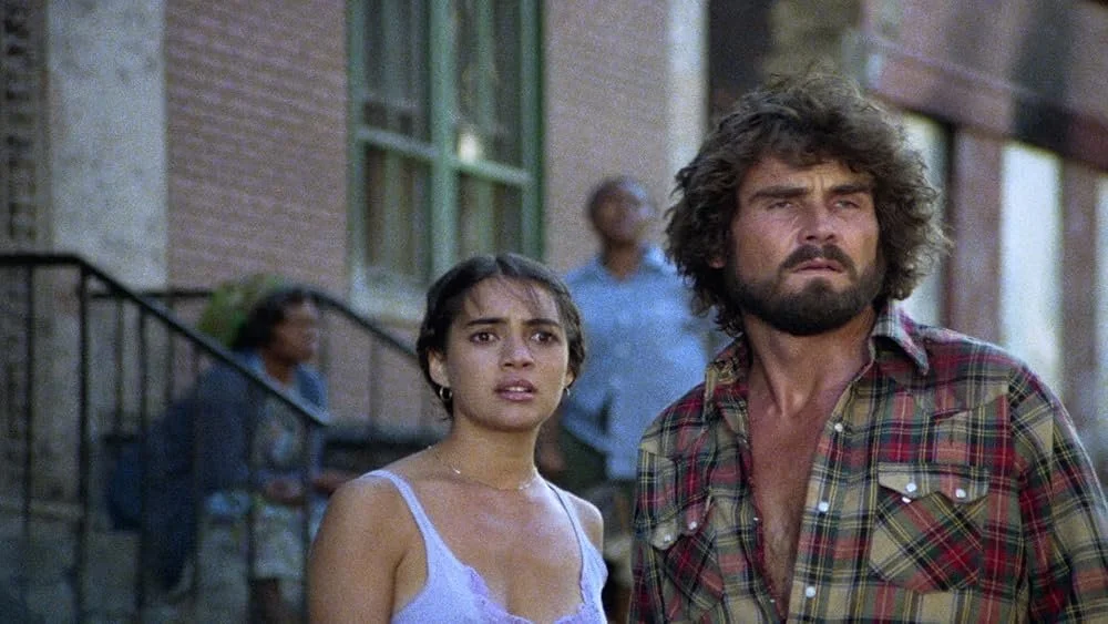 A young woman in a lavender camisole and a man with disheveled curly brown hair and short beard wearing a partially unbuttoned plaid shirt stand side by side in front of city buildings with concerned looks on their faces.