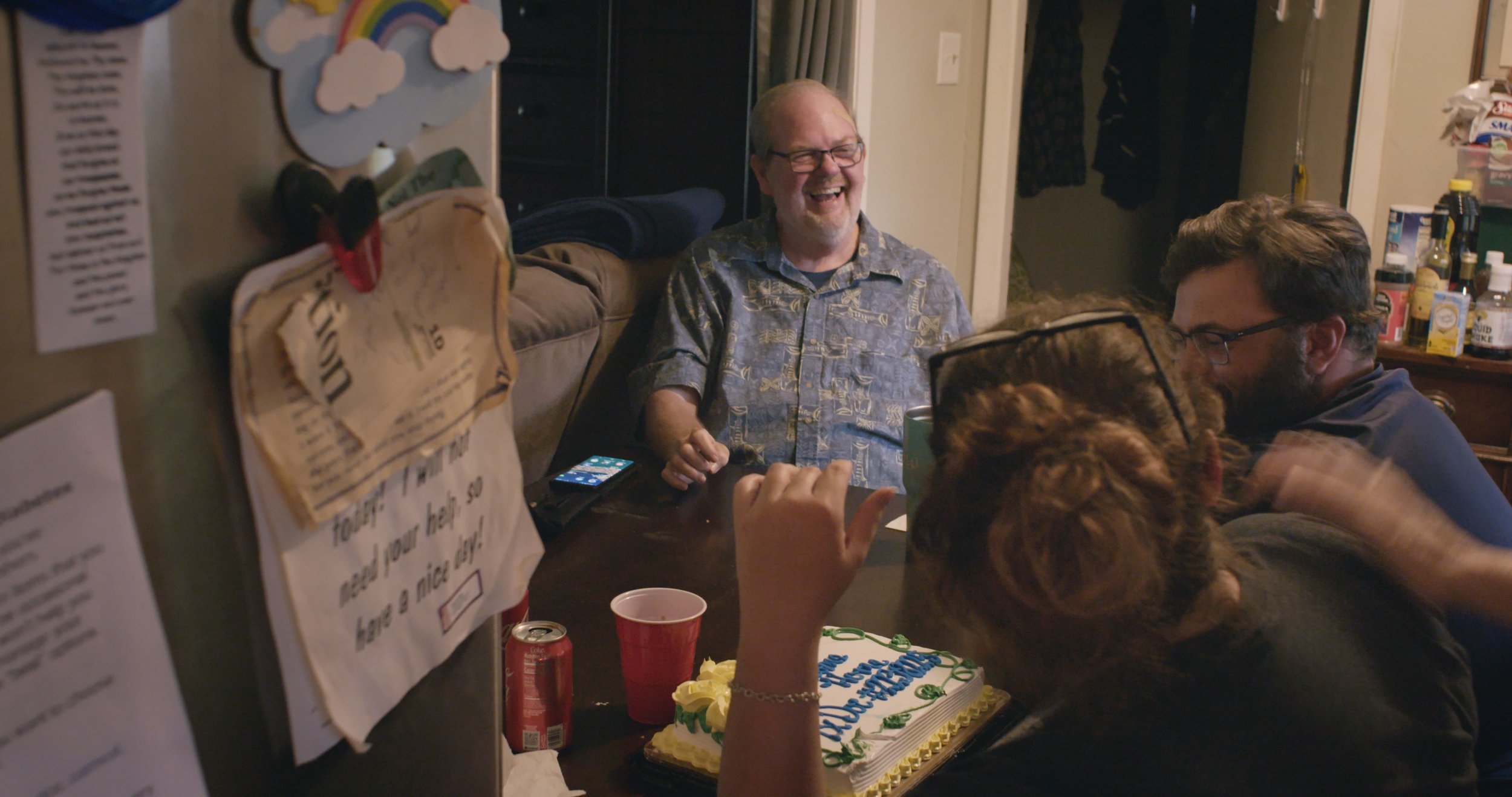 Walter, Leslie, and Leslie's husband sit at a kitchen table laughing. There is a cake sitting in front of Leslie.