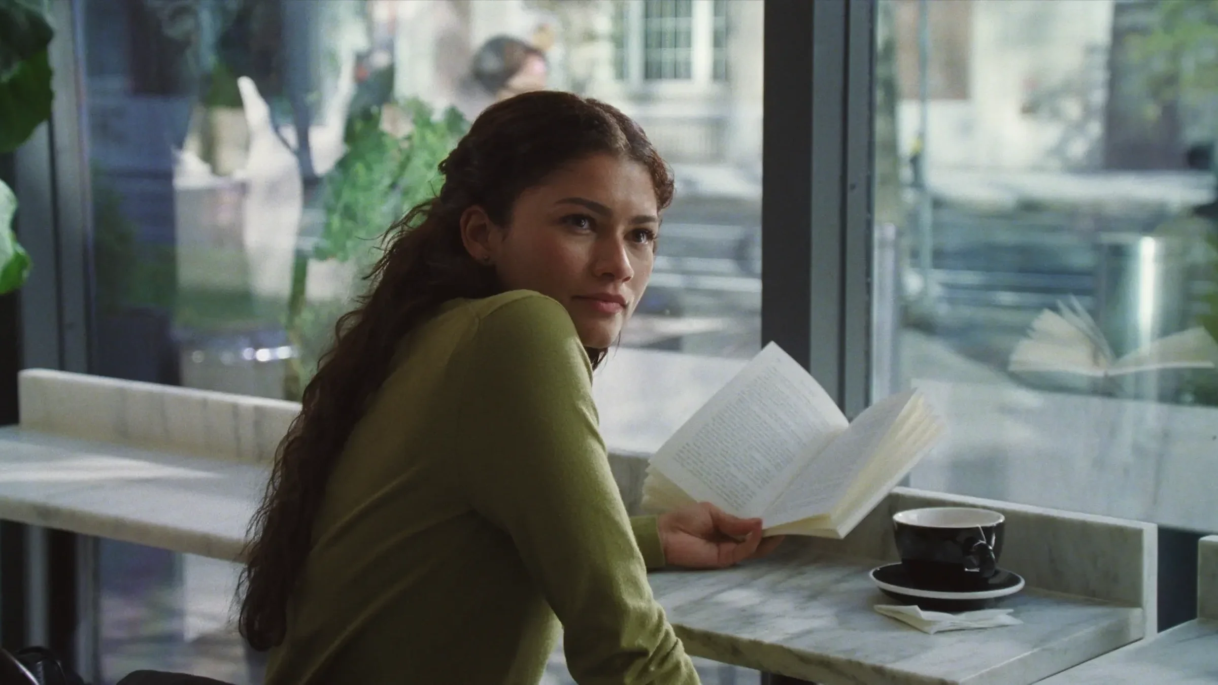 Emma (played by Zendaya) looks towards the camera with a book in her hand while sitting in a coffee shop in the film, 'The Drama.'