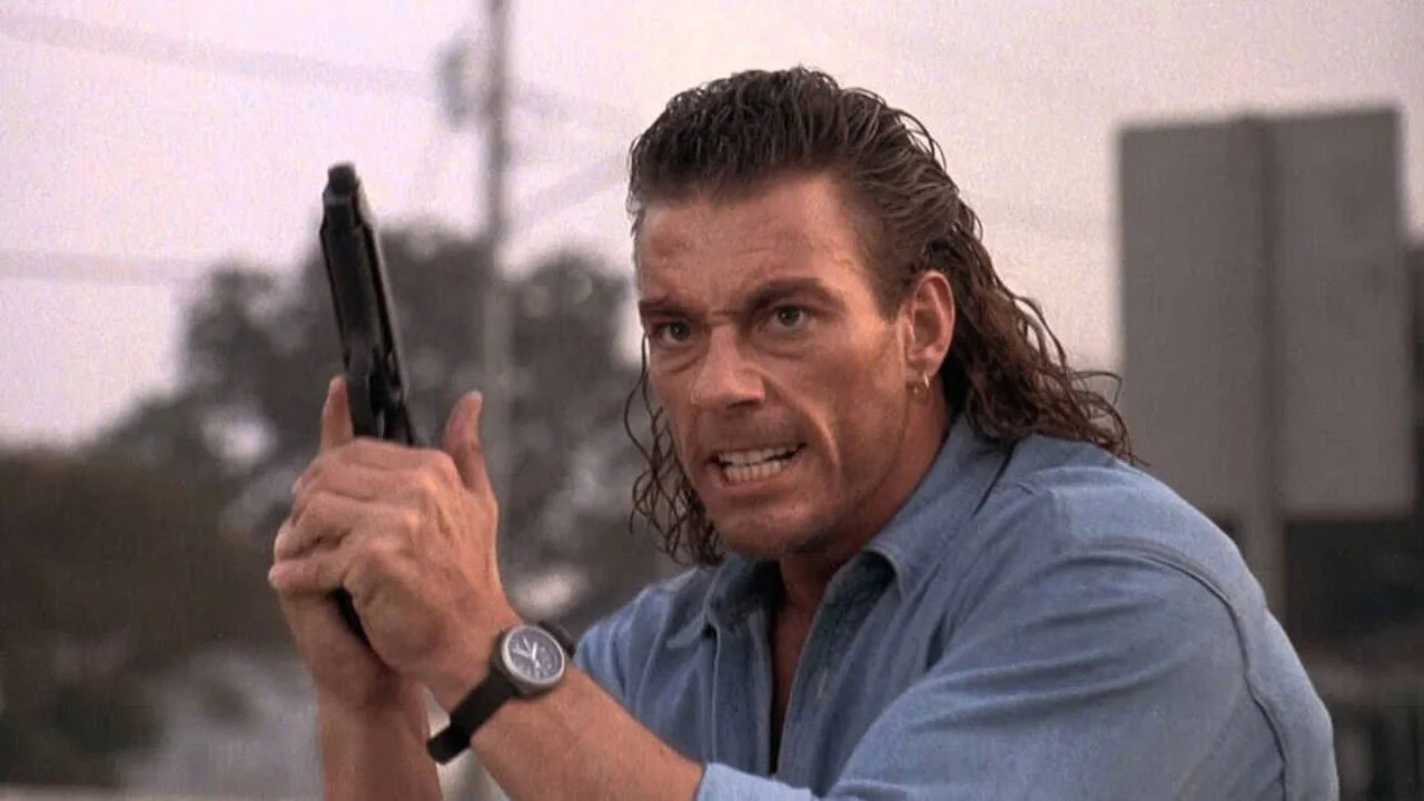 Jean Claude Van Damme grimaces angrily and holds a handgun, looking ready to fire.