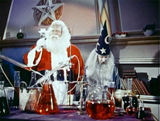 Santa stands next to Merlin in a potions lab, behind a table covered in bottles filled with red liquid.