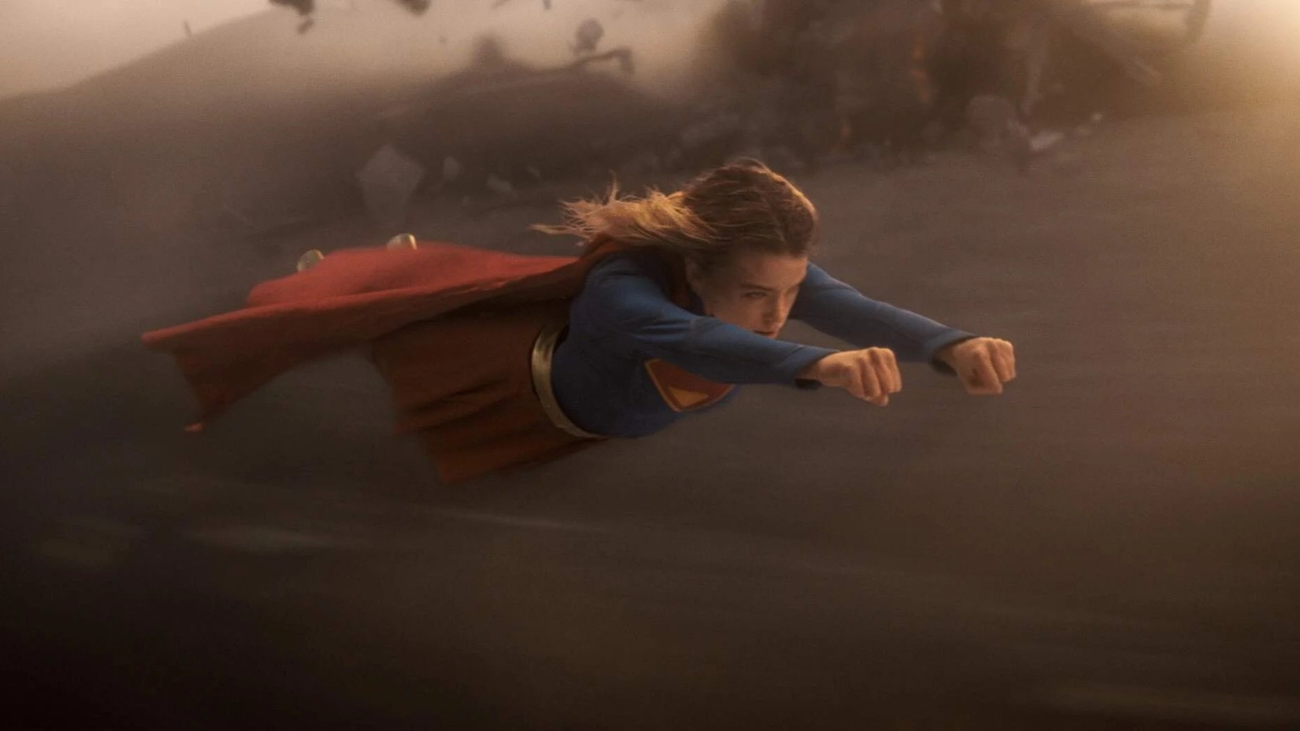 Supergirl (played by Milly Alcock) soars through a rocky planet in the classic Superman pose.