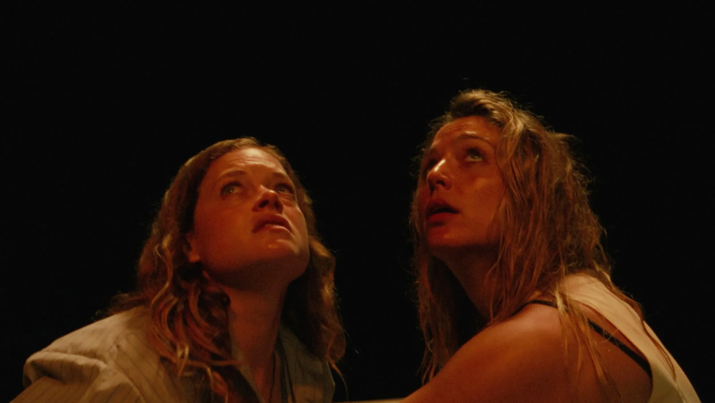 Two women, basked in reddish light against a pitch-black sky, looking upward with fearful expressions.