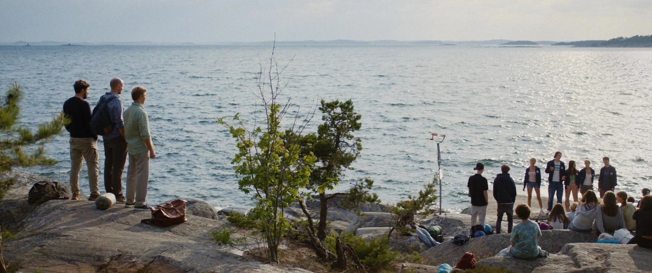 Three men stand on a rock overlooking a beach, where a group of children and camp counselors stand.
