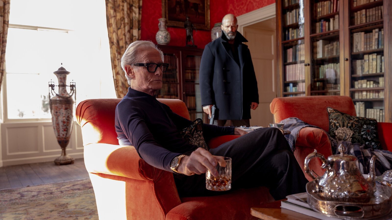 In a large, expensive looking room, Bill Nighy sits holding a glass of whiskey as Jason Statham looms in the background in Shelter.