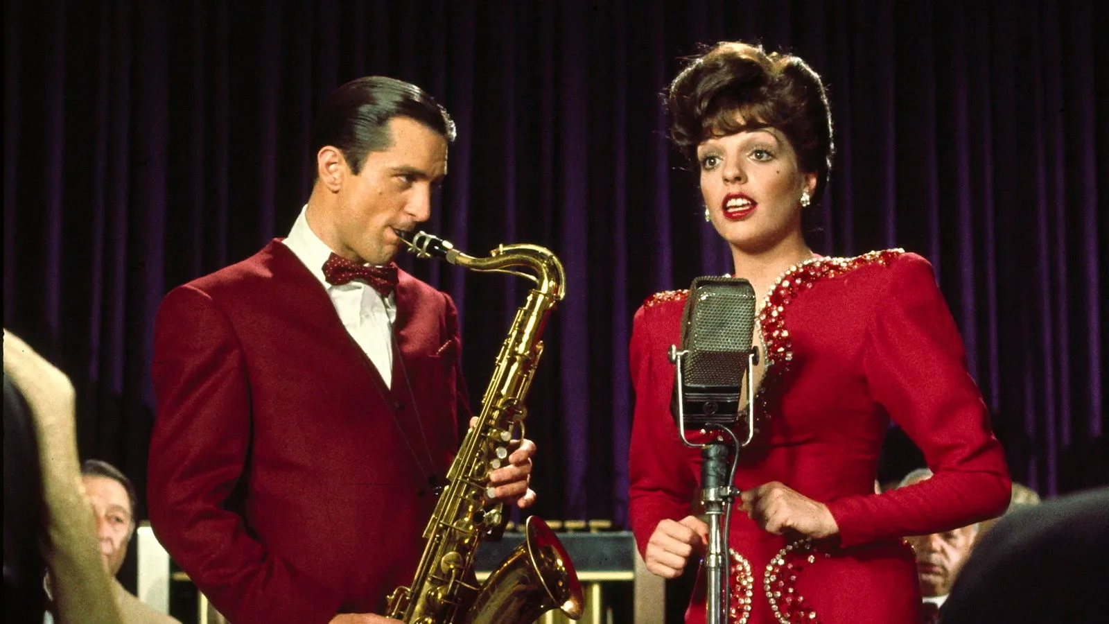 Robert De Niro, wearing a maroon suit and matching bow tie, plays a saxophone while Liza Minnelli, wearing a red dress with sequin details, sings into a microphone. They stand on a stage in front of a deep purple velvet curtain.