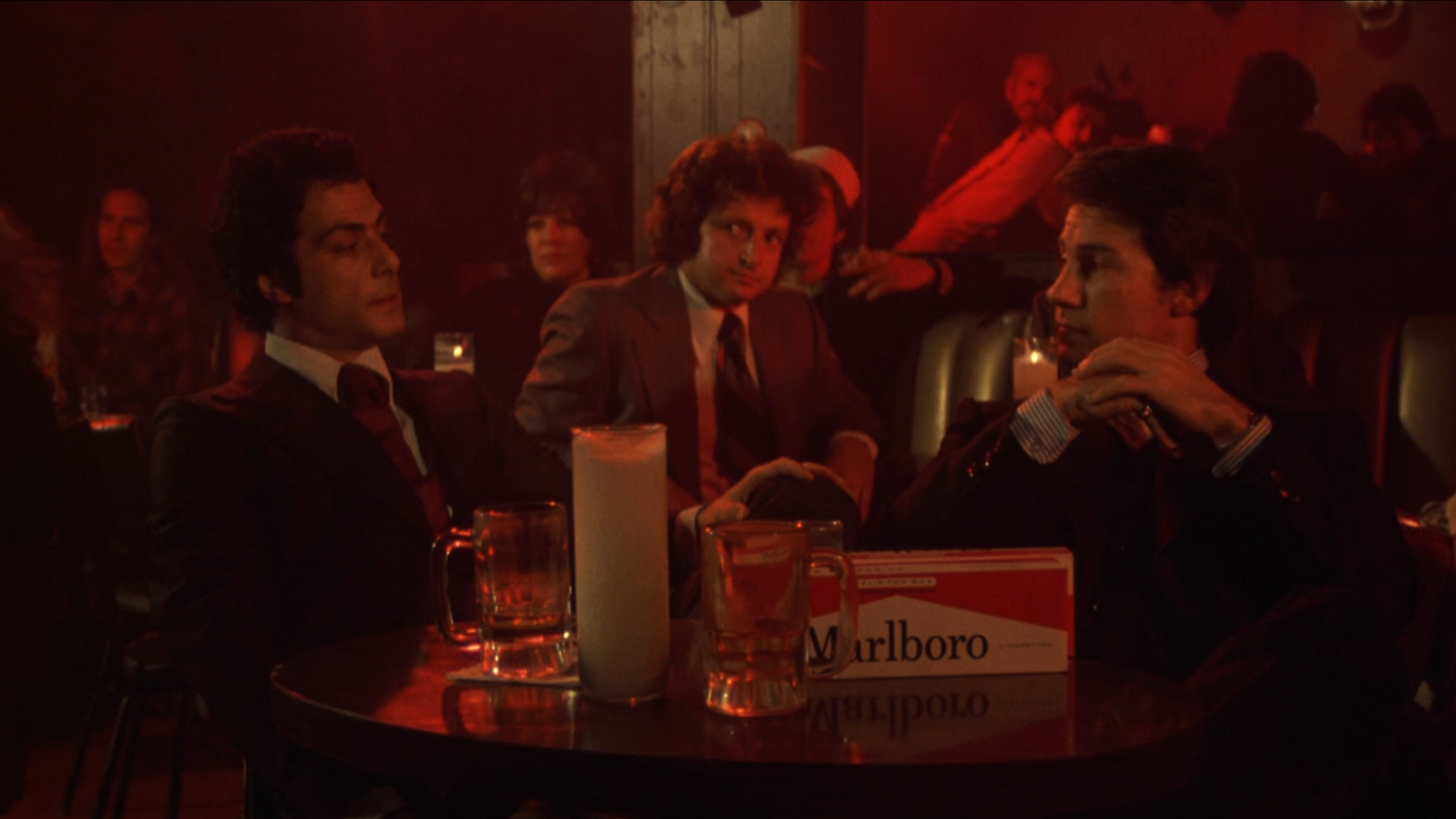 Richard Romanus and Harvey Keitel look at each other over a table in a low-lit bar.
