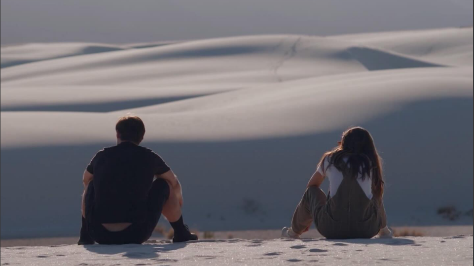 Phil Dunster as Rex sits on a sand dune next to Chloe Bennet's Minnie in Hello Out There.