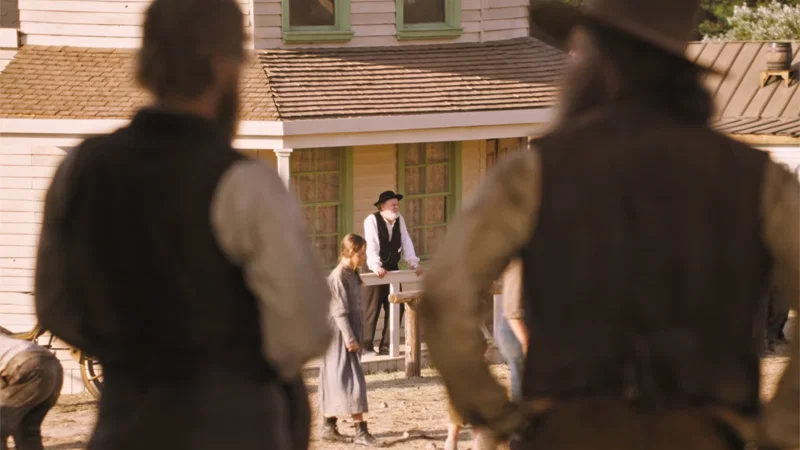 Two men standing in the foreground with their backs to the camera observe a man and a woman in front of a house