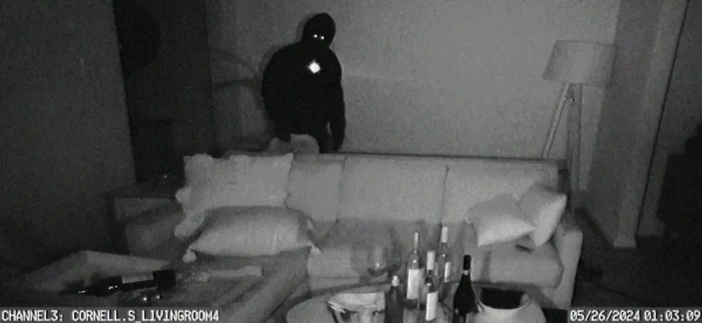 A frame from a black-and-white nighttime security camera showing a shadowy figure with two glowing, white eyes standing behind a living room sofa.