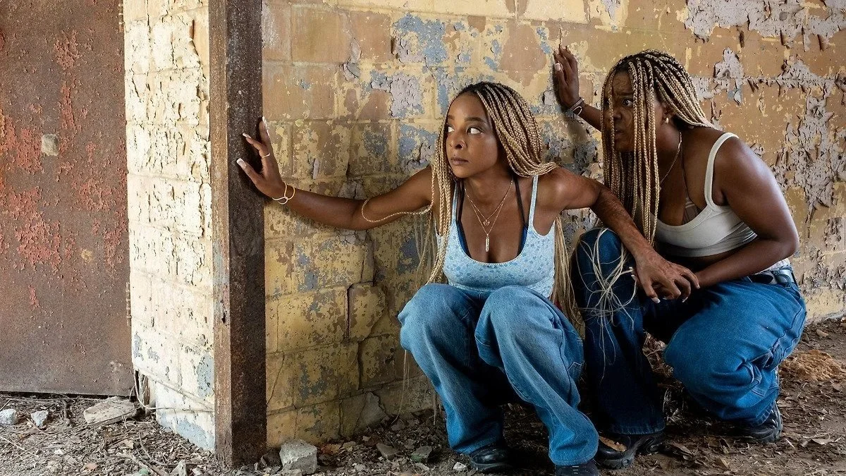 Kara Young as Racine squats next to Mallori Johnson as Anaia in front of a peeling brick wall in the film Is God Is.
