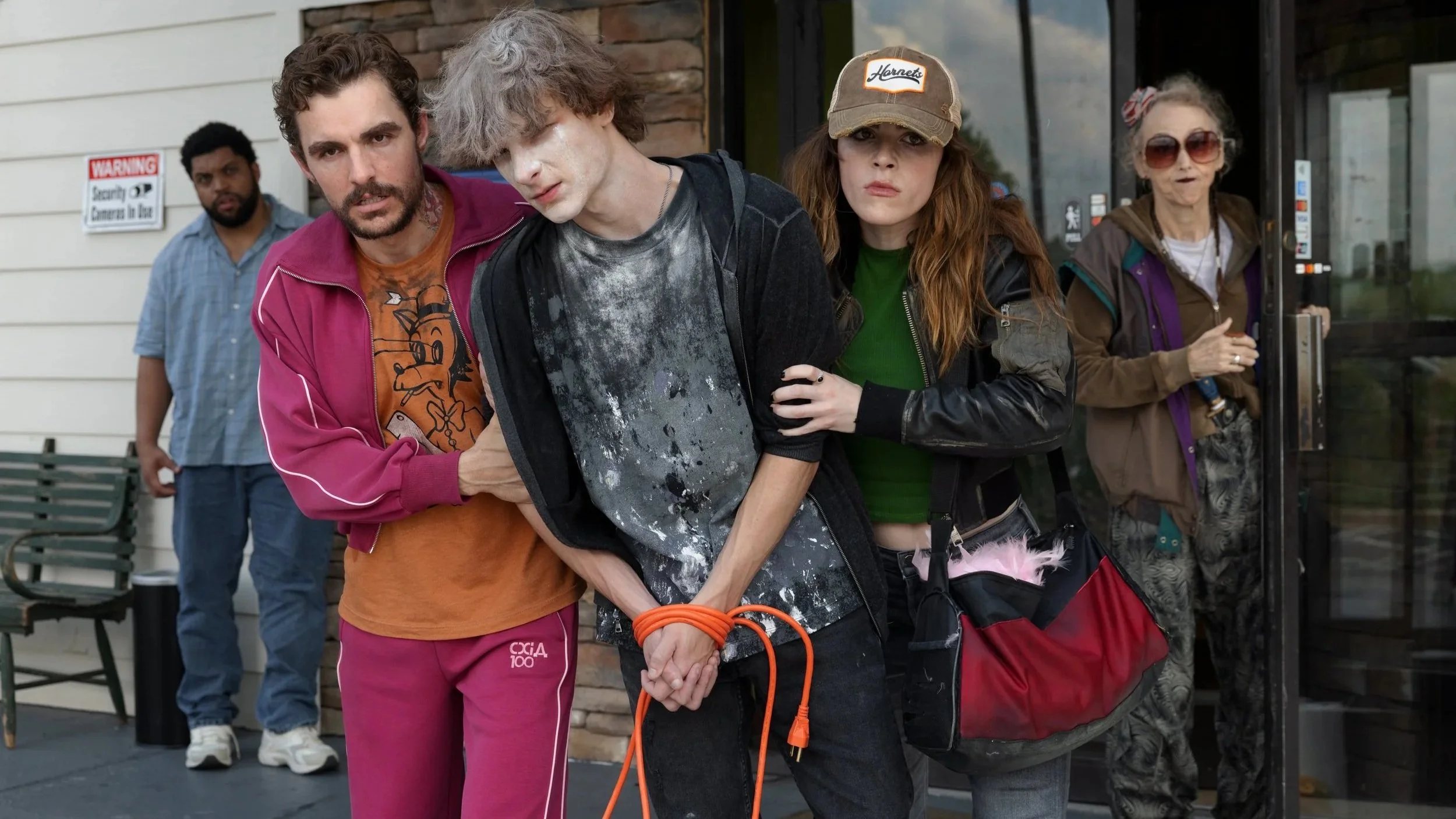 Davis (O'Shea Jackson Jr.) stands in the background as Mark (Dave Franco) and Irina (Kiernan Shipka) haul Sheridan (Mason Thames) somewhere in the film Idiots.
