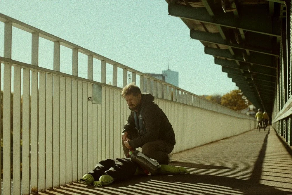A man with short hair and a beard wearing a black windbreaker kneels beside the handrails of a bridge, looking pensive.