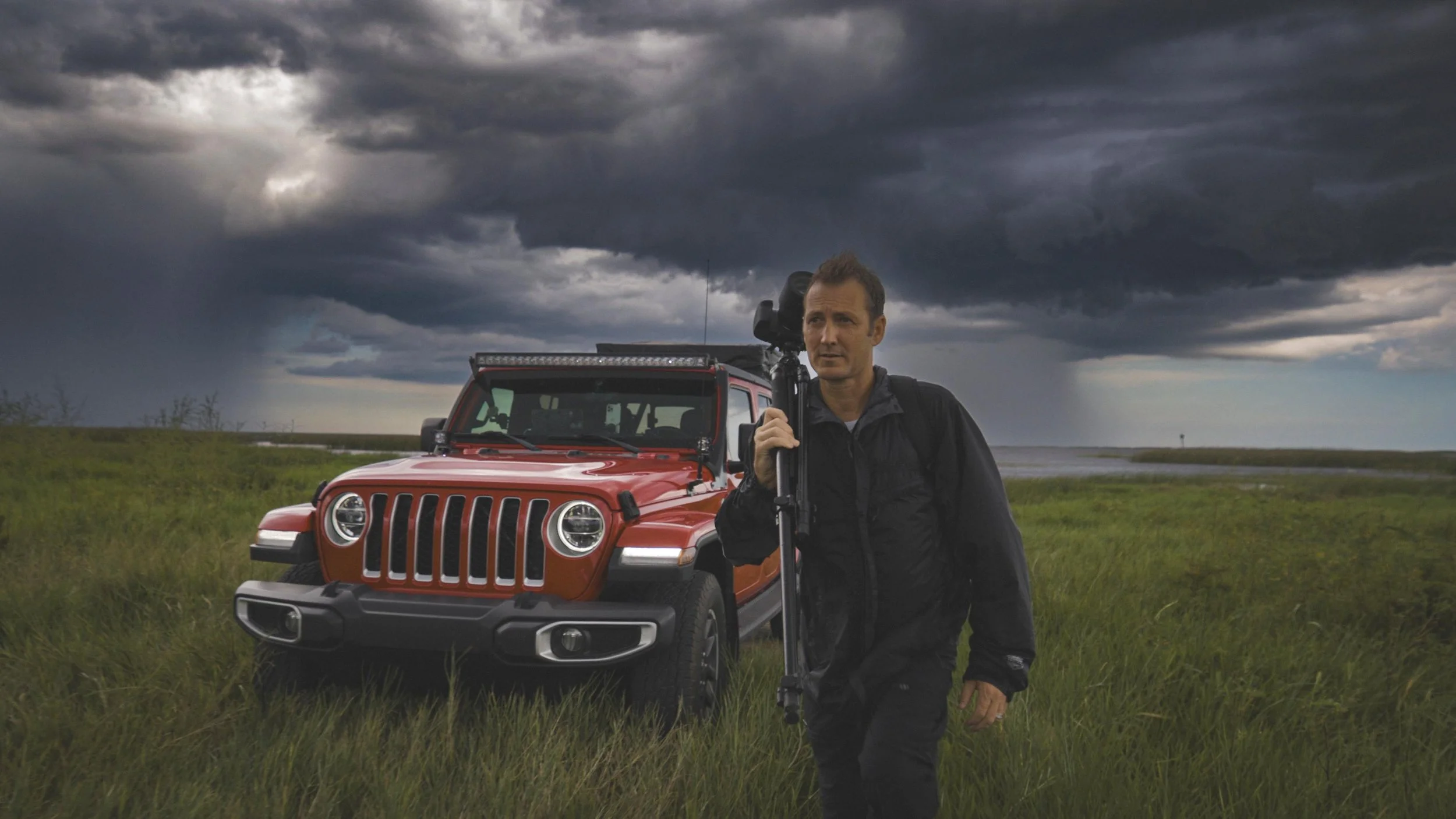 Jeff Gammons stands holding a camera in an open field. Grey storm clouds fill the sky behind him.