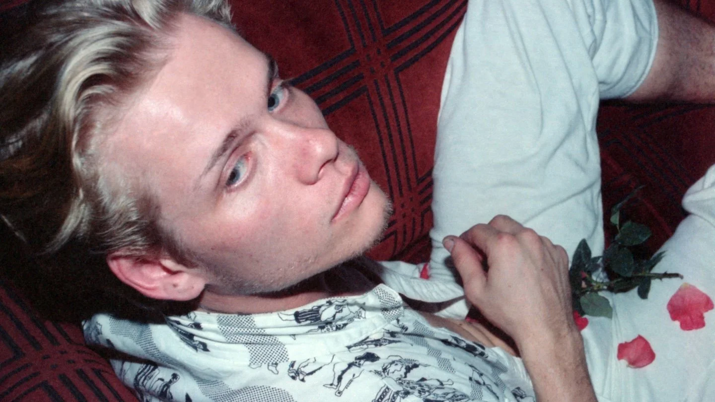 Billy London, a white man with blonde hair, blue eyes, and some facial scruff, sits reclined near the camera wearing all white and holding a rose
