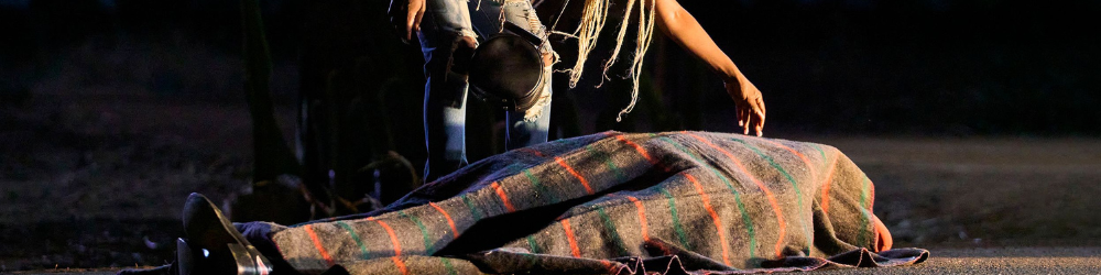 A woman, her top half cut out of the frame, bends over a covered body in the road in On Becoming a Guinea Fowl.