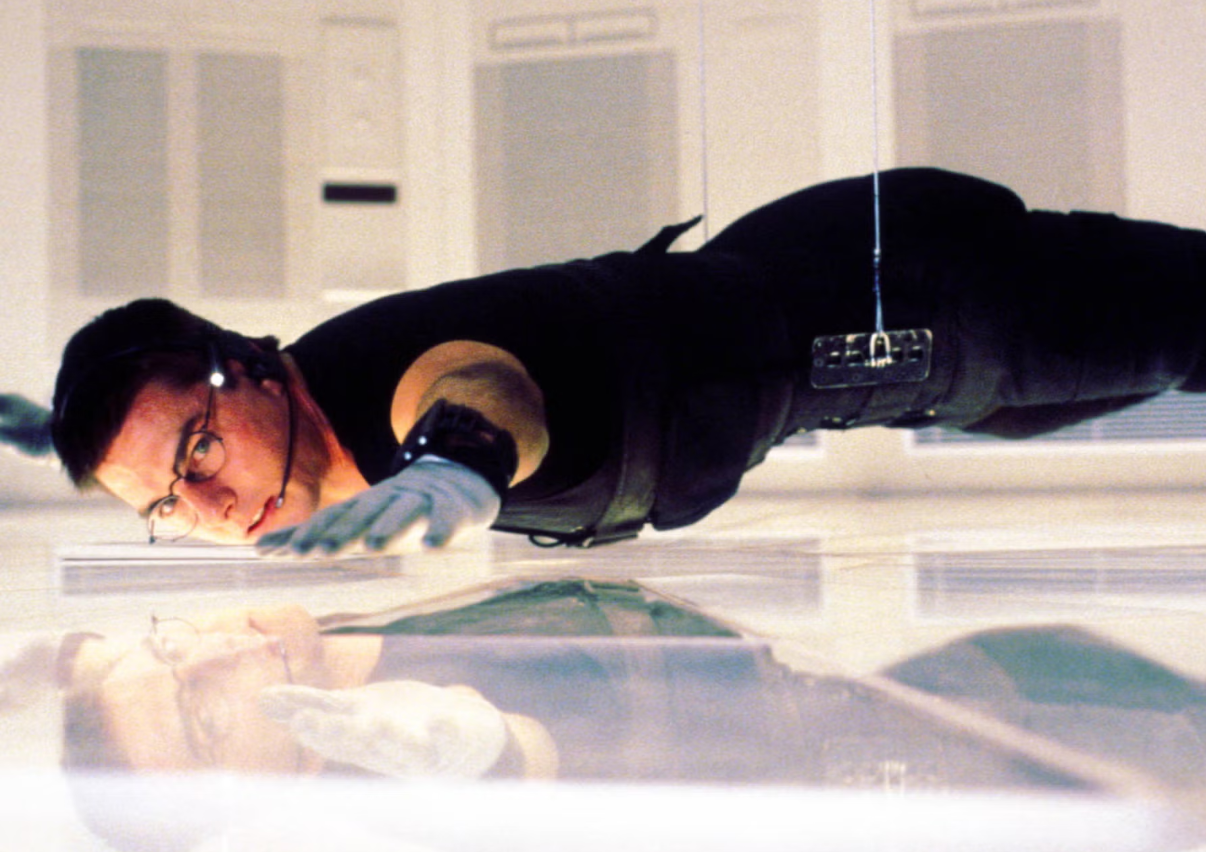 Tom Cruise, dressed in all black and wearing glasses and a headset microphone, suspended by a harness inches from a shiny and reflective white floor.